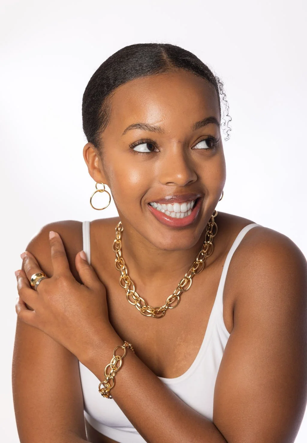 A smiling woman wearing gold jewelry, including a necklace, bracelet, rings, and hoop earrings, against a plain white background.
