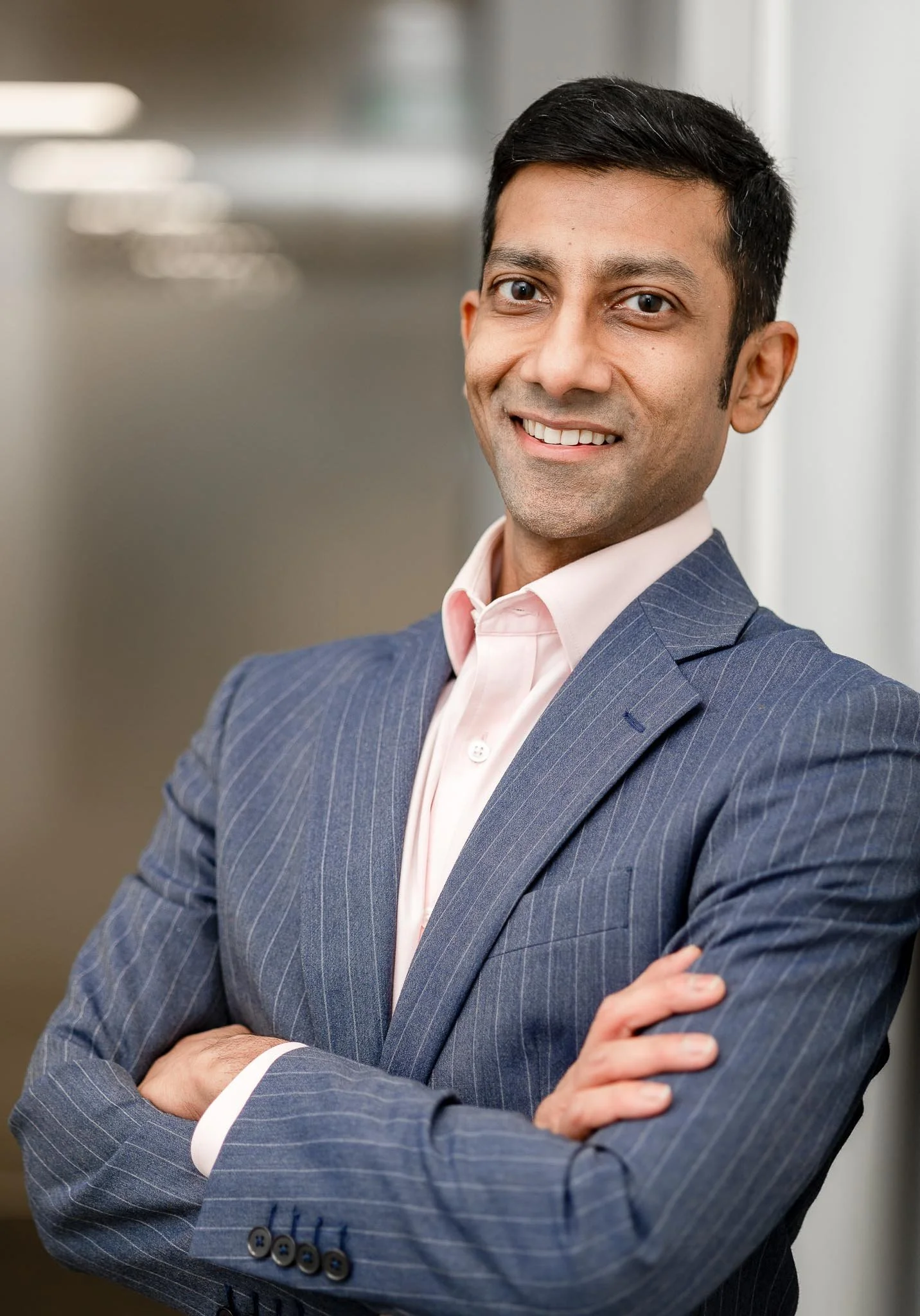 A confident man in a navy pinstripe suit with arms crossed, smiling in an office setting.