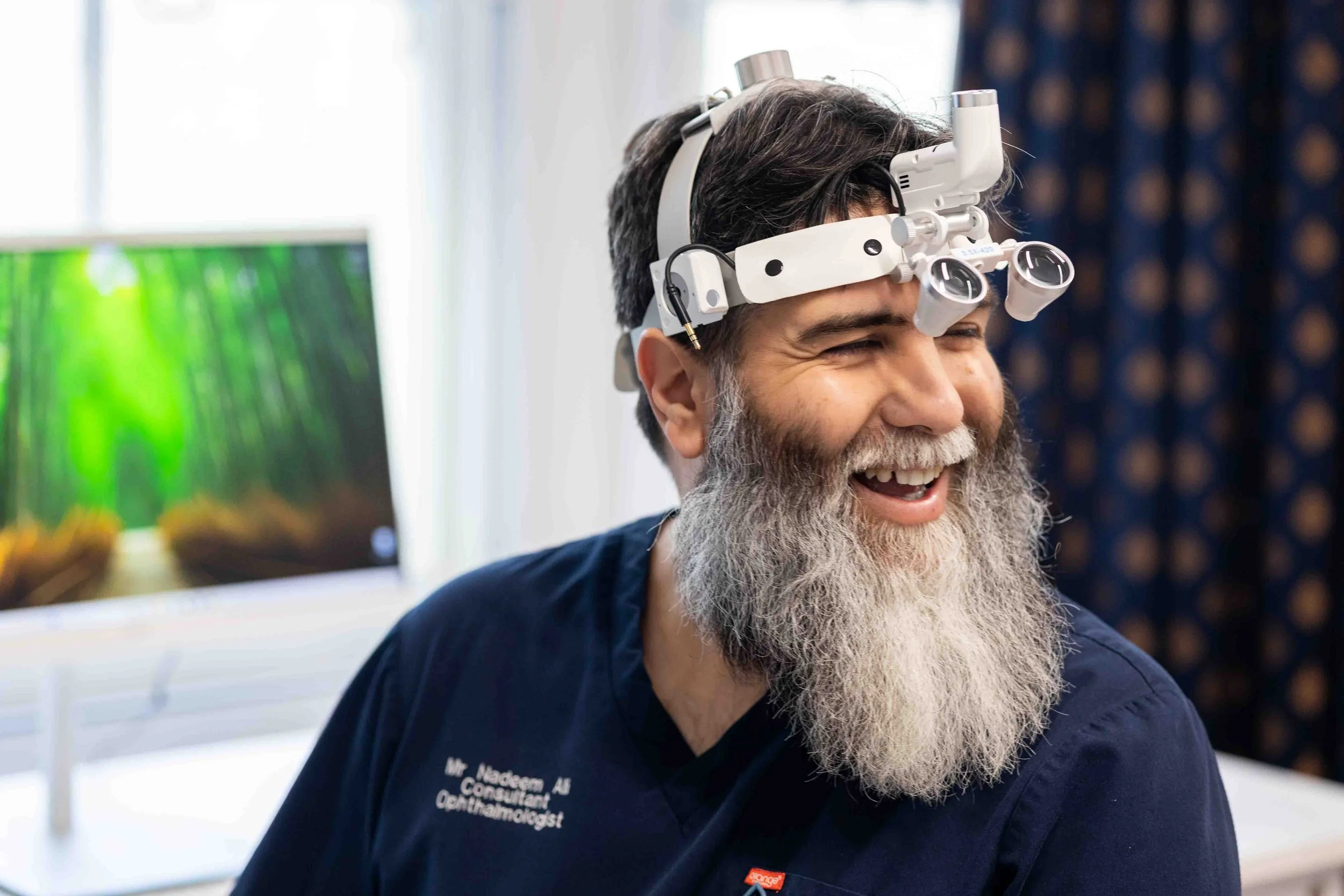 A smiling man with a gray beard wearing a headlamp or magnifier with lenses, sitting in a well-lit room with a computer monitor in the background.