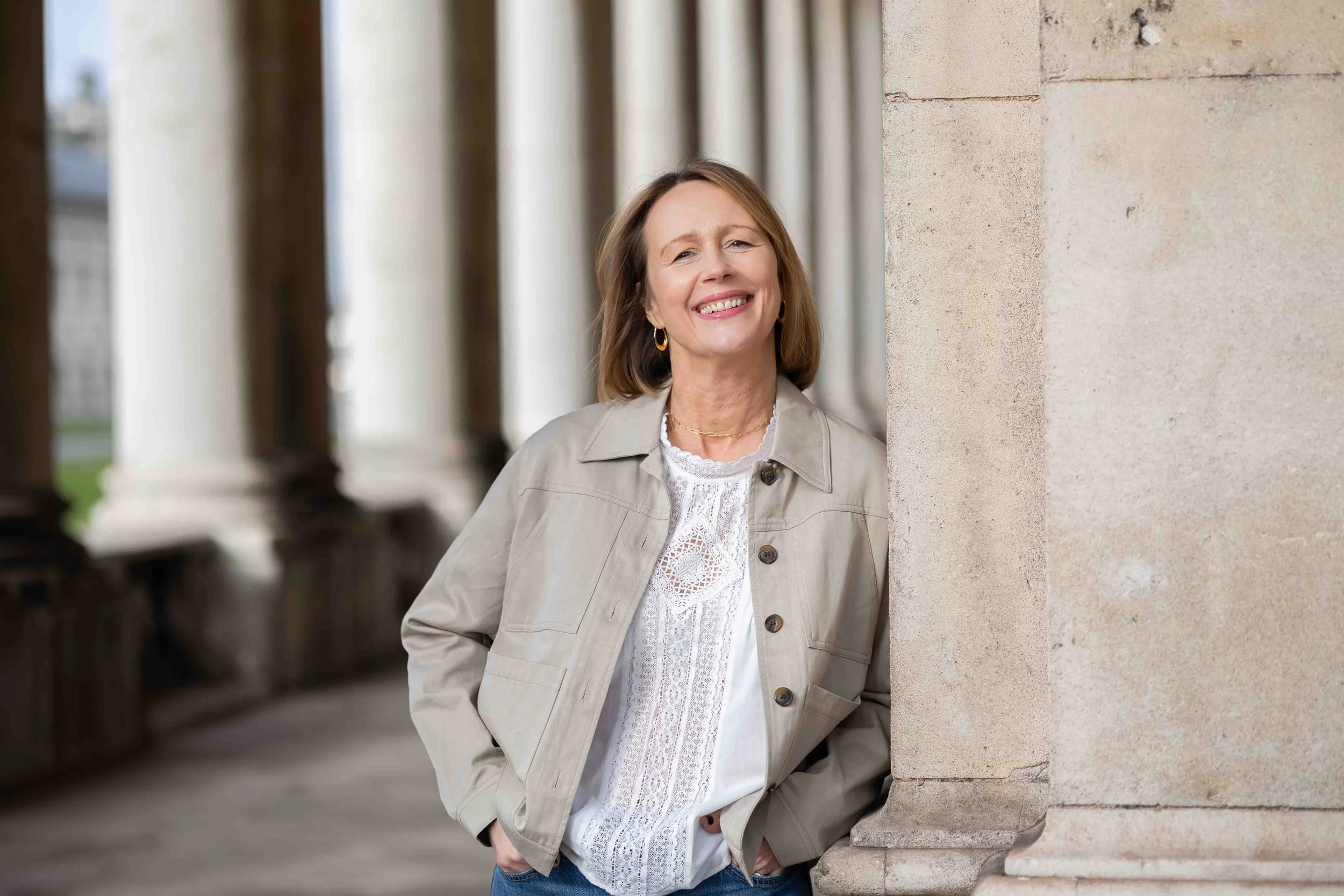 A woman with shoulder-length light brown hair, wearing a beige jacket and white lace blouse, leaning against a stone column outdoors, smiling at the camera.
