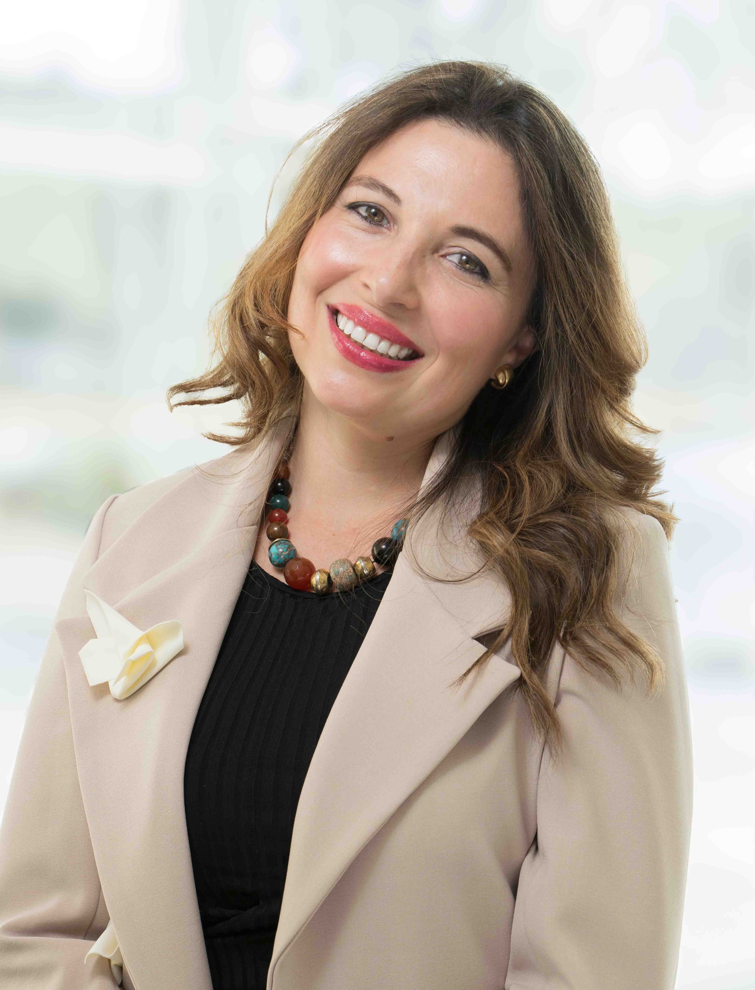 A woman with wavy brown hair wearing a beige blazer, black top, colorful beaded necklace, gold earrings, and a white flower accessory on her blazer, smiling confidently.