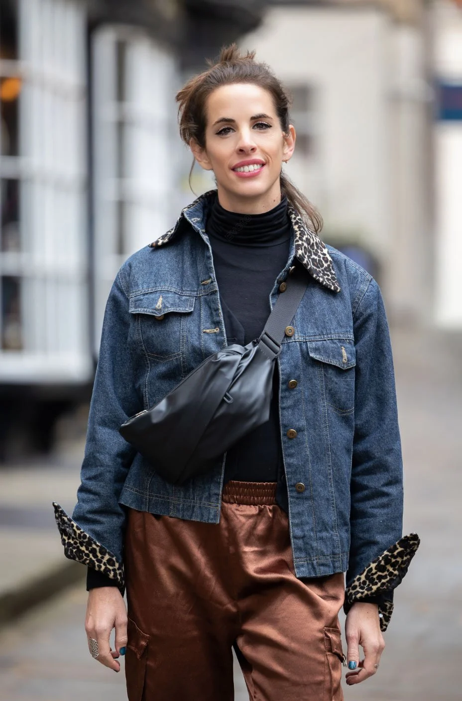 A young woman with brown hair tied back, smiling, wearing a denim jacket with leopard print collar and cuffs, a black turtleneck, and brown pants, walking outdoors.