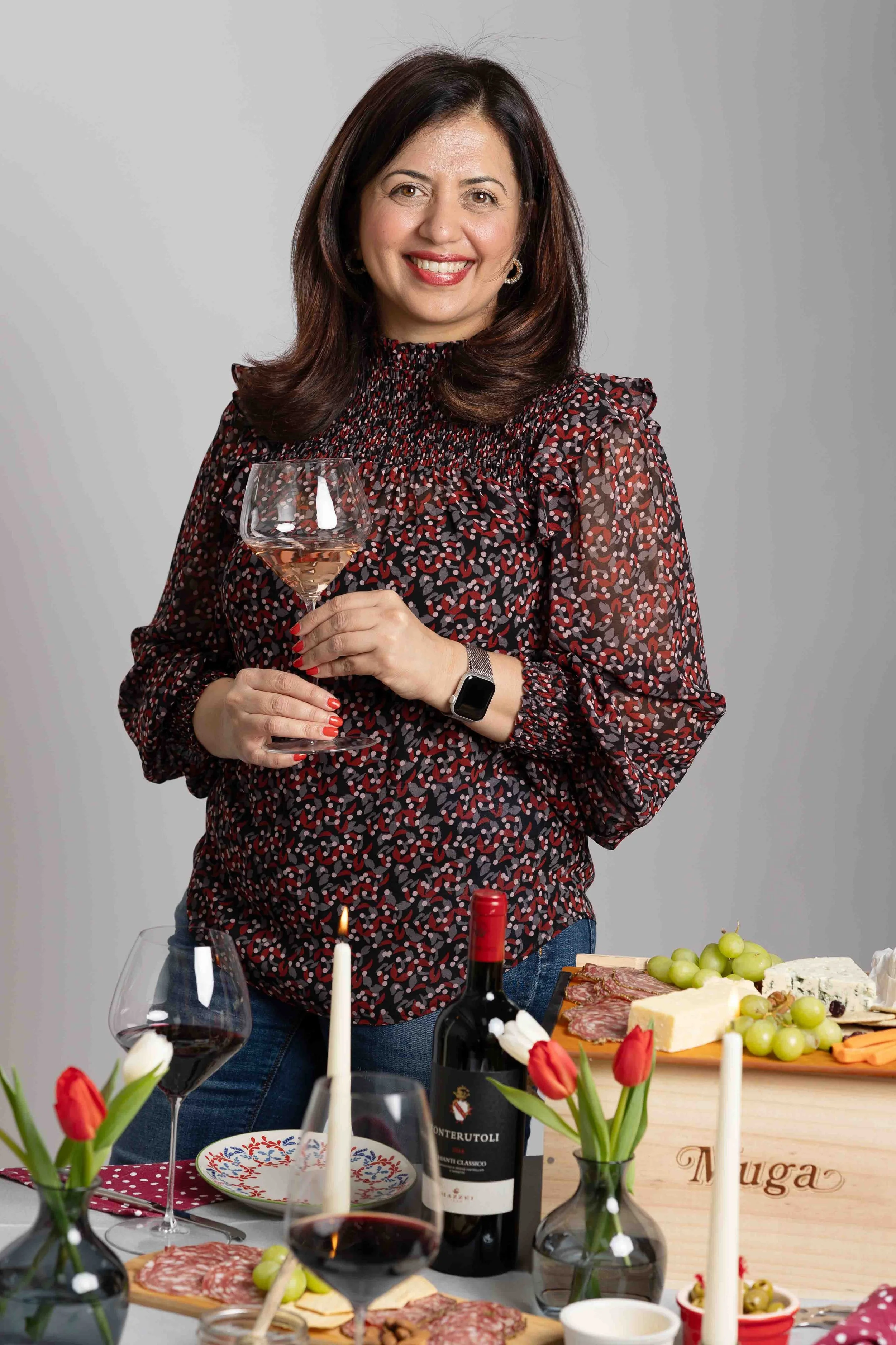 A woman with brown hair wearing a red patterned blouse holding a glass of rosé wine at a table set for a celebration, with food, wine, candles, and flowers.