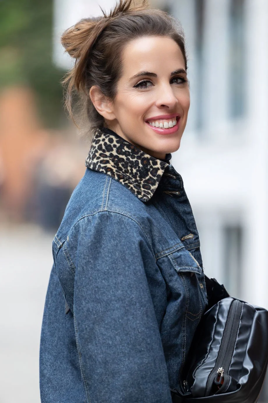 A woman smiling, wearing a denim jacket with a leopard print collar, holding a black bag, outdoors with blurred background.