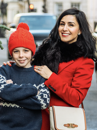 A woman and a young girl posing together outdoors on a city street, both smiling. The woman has long dark hair and is wearing a red coat, while the girl is wearing a blue sweater with a winter pattern and a red knit hat.
