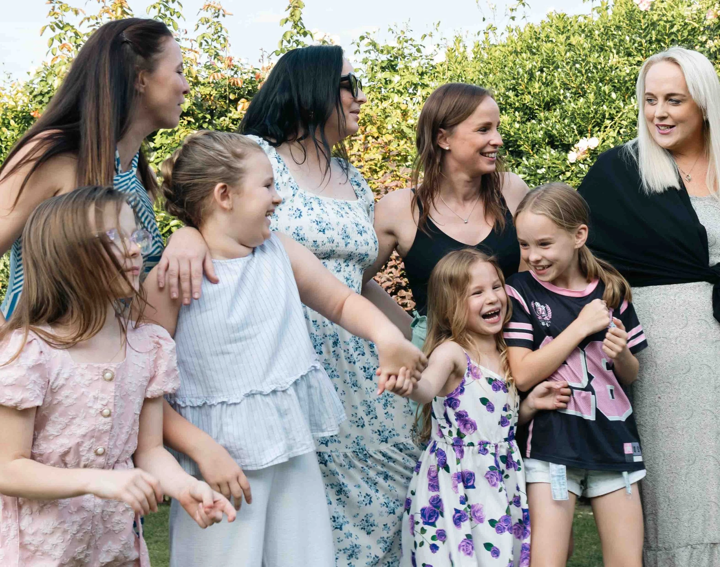 Group of women and young girls smiling and holding hands in a garden with green bushes in the background.