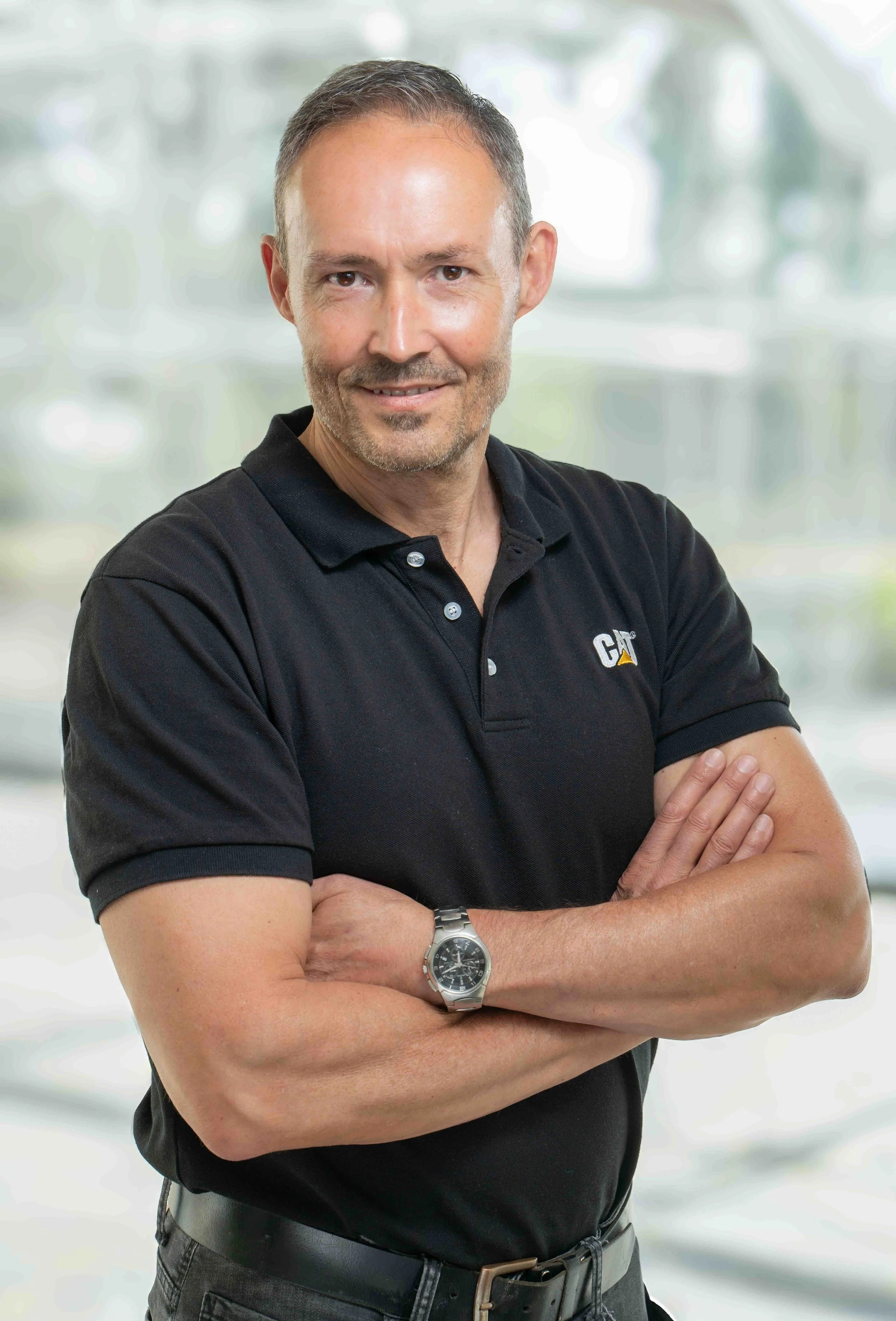 A middle-aged man with short gray hair and stubble, wearing a black collared shirt with a small logo, standing with arms crossed and smiling, in front of a blurred background.