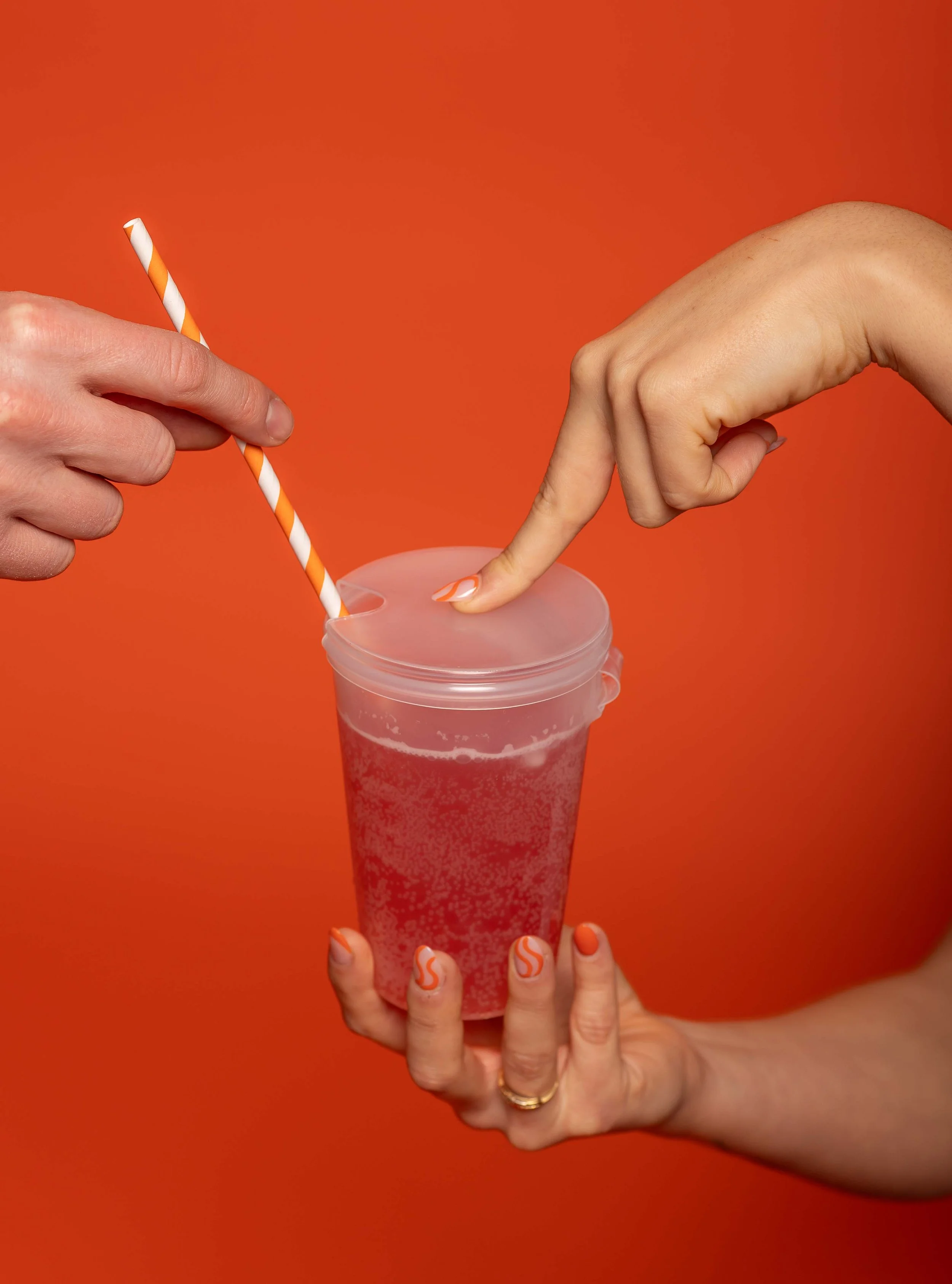 Two hands with orange and white painted nails interact with a pink drink in a clear cup with a lid, a striped straw, and a bright orange background.
