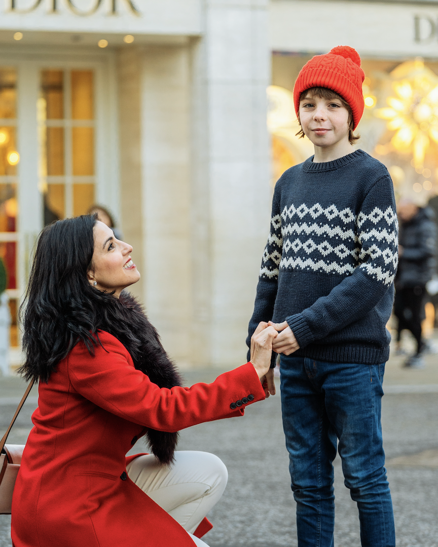 A woman in a red coat kneels and holds hands with a young boy wearing a red knit hat and a blue sweater with white geometric patterns, outside in a city street during the winter holiday season.