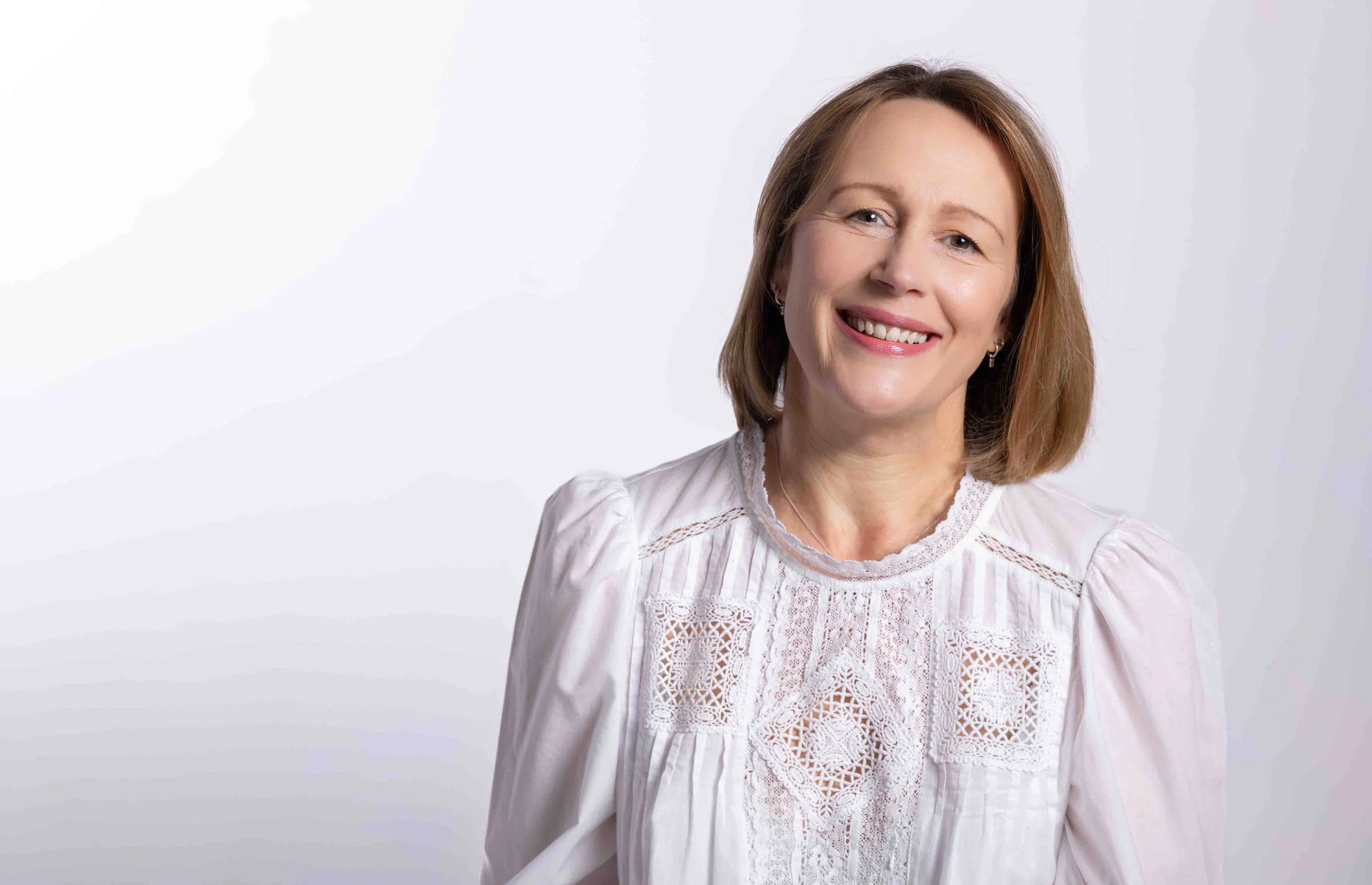 A smiling woman with shoulder-length brown hair, wearing a white lace blouse, standing against a plain white background.