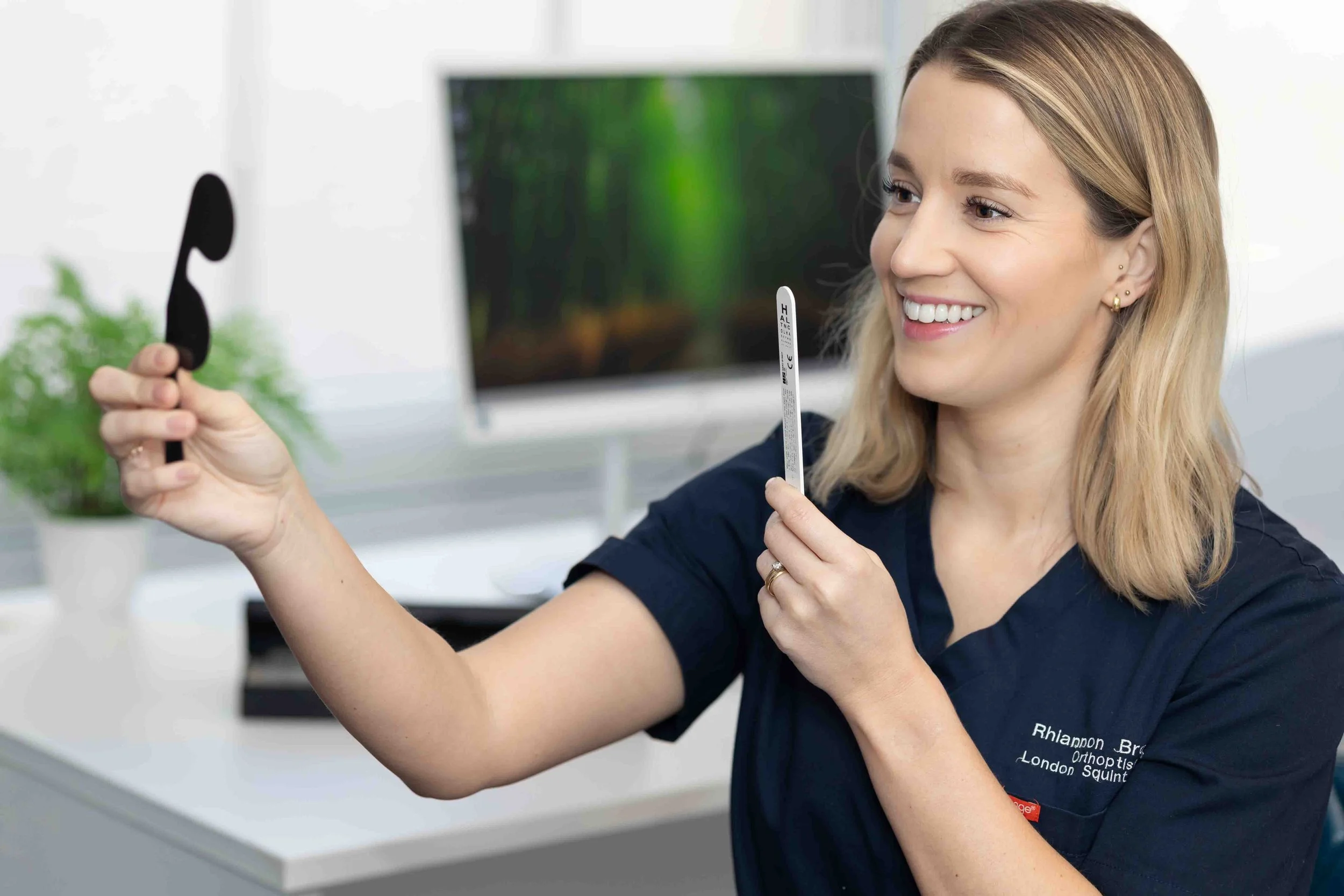 A smiling woman in navy scrubs holding an eye patch and an eye measurement tool in a medical office.