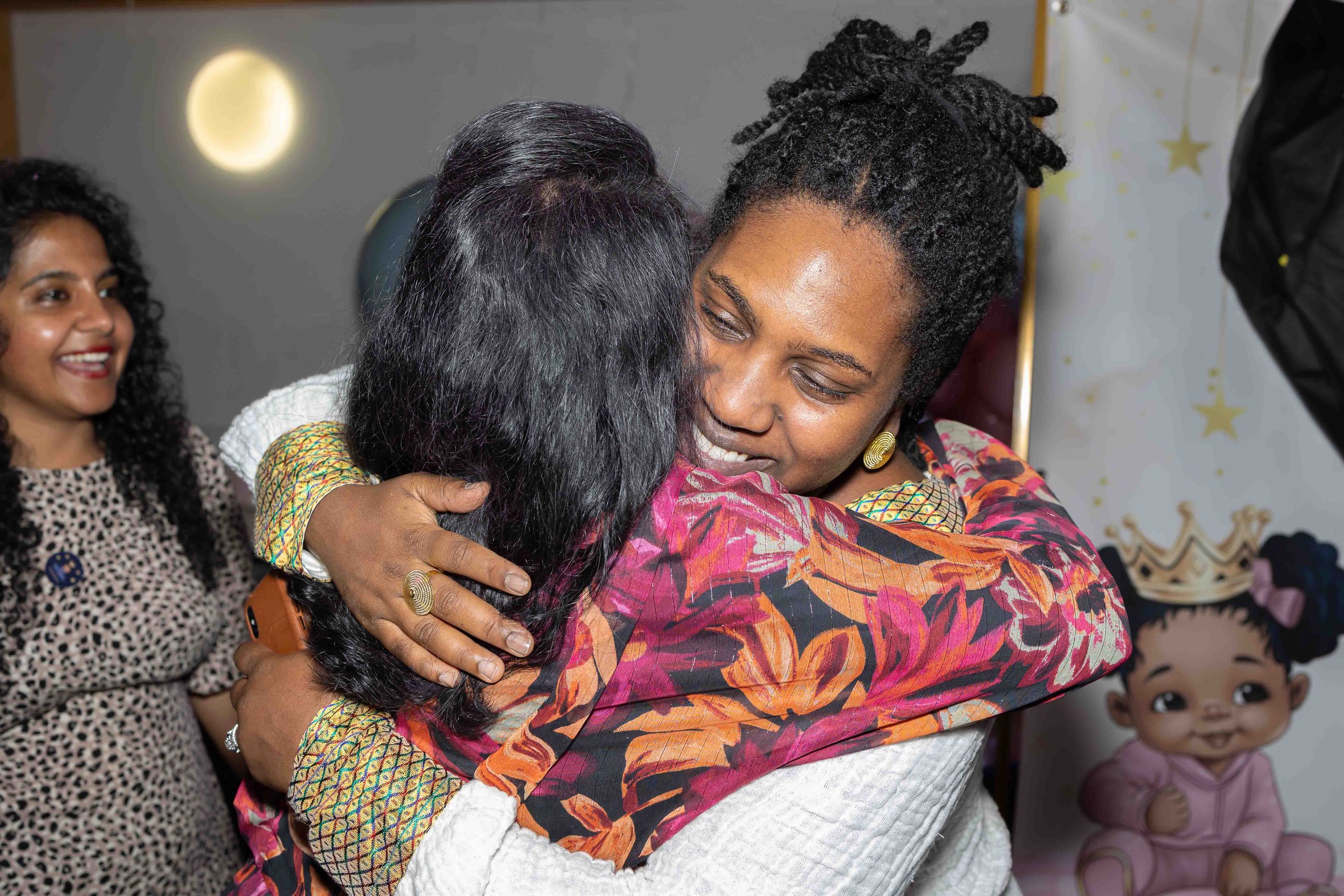 Two women hugging warmly, with a third woman smiling in the background, at a celebration or event with decorative balloons and wall art.