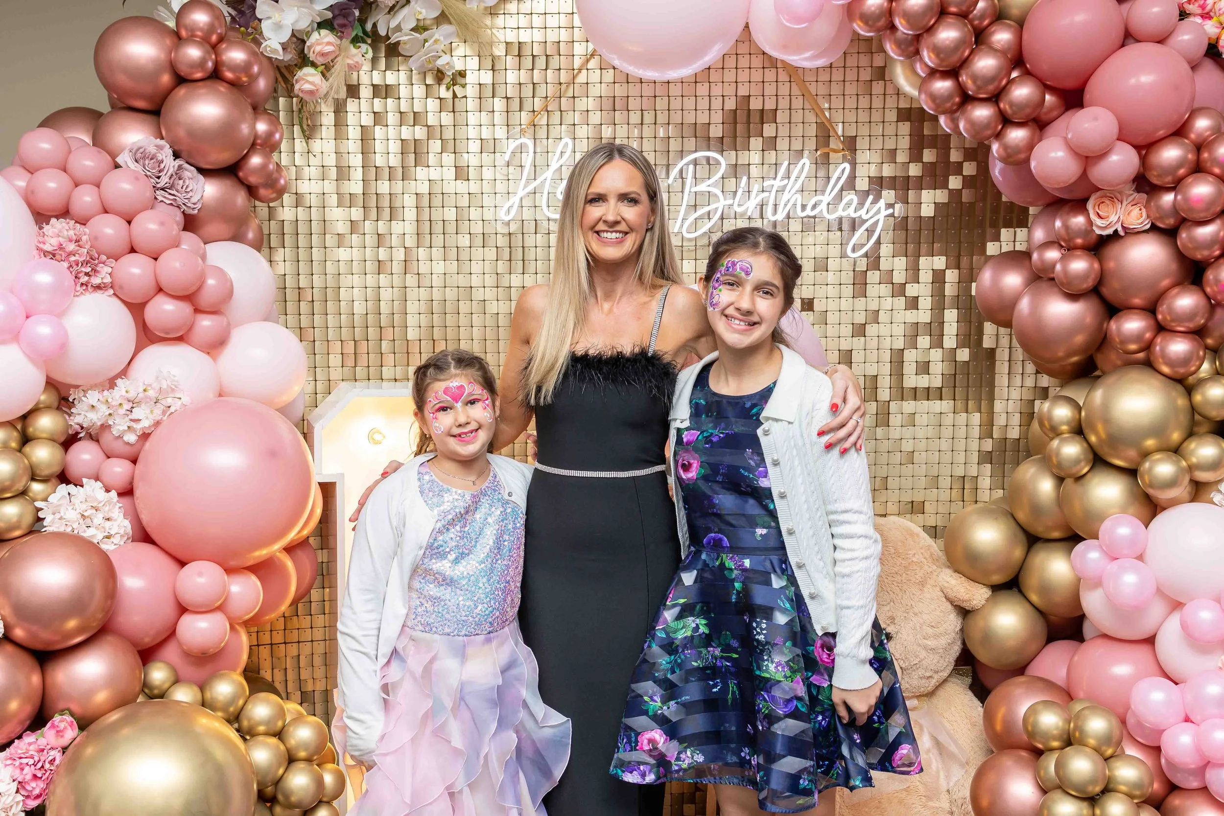 A woman and two young girls smiling at a birthday celebration with pink, gold, and white balloons and flowers, and a sign that says 'Happy Birthday' in the background.