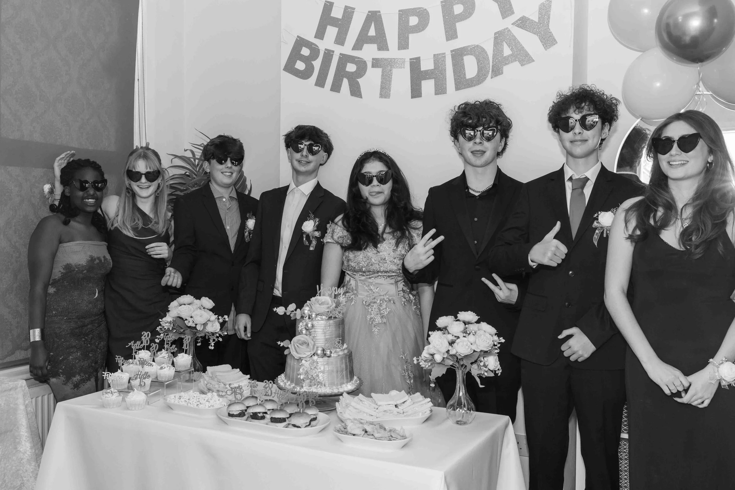 Group of people celebrating a birthday at a party, dressed in formal attire with sunglasses, standing behind a table with cake, cupcakes, and snacks, in front of a 'Happy Birthday' banner and balloons.