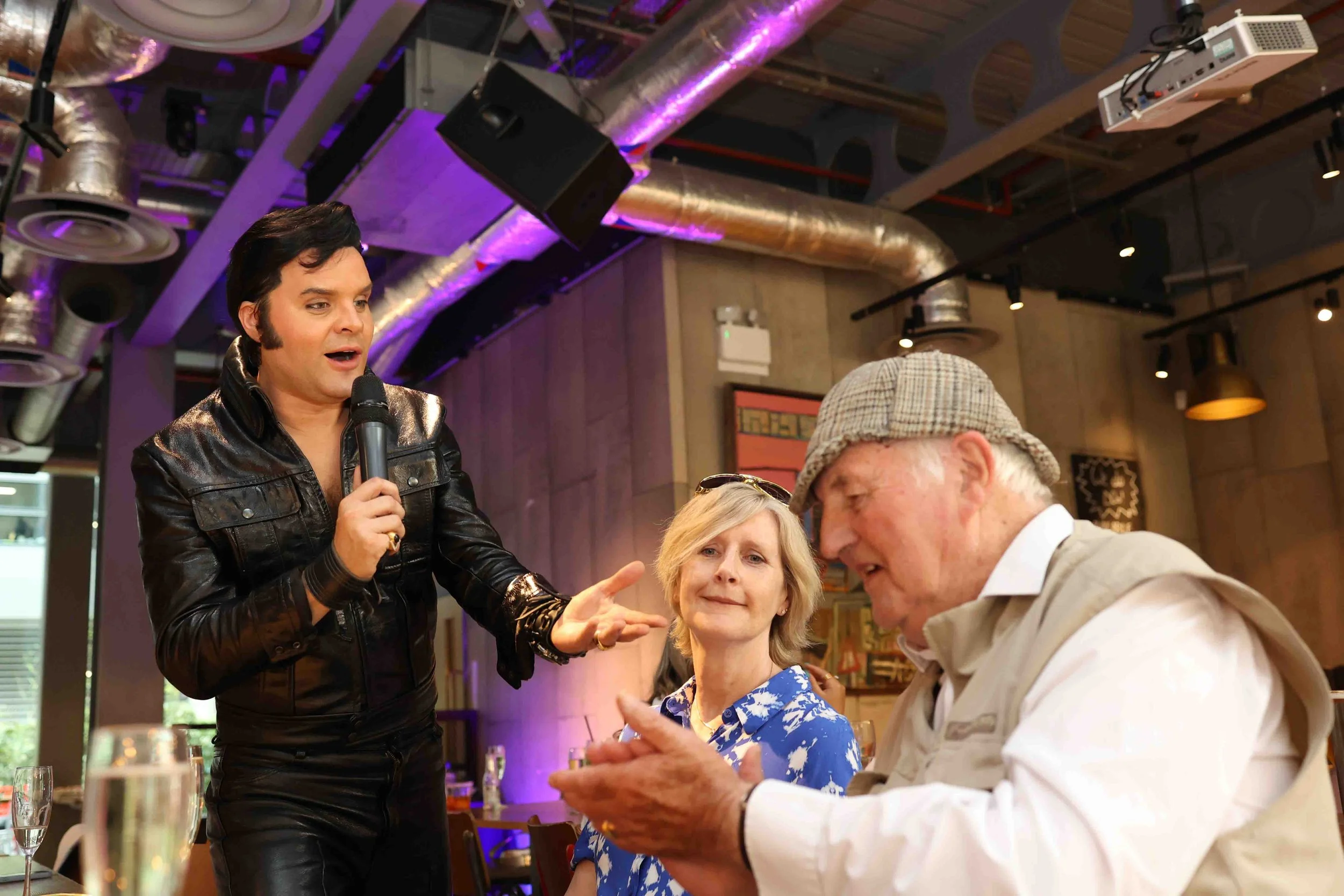 A man dressed as Elvis Presley performing at a karaoke night in a restaurant, singing into a microphone while pointing at an elderly man in a cap. An elderly woman sitting between them looks on, with a glass of champagne on the table.