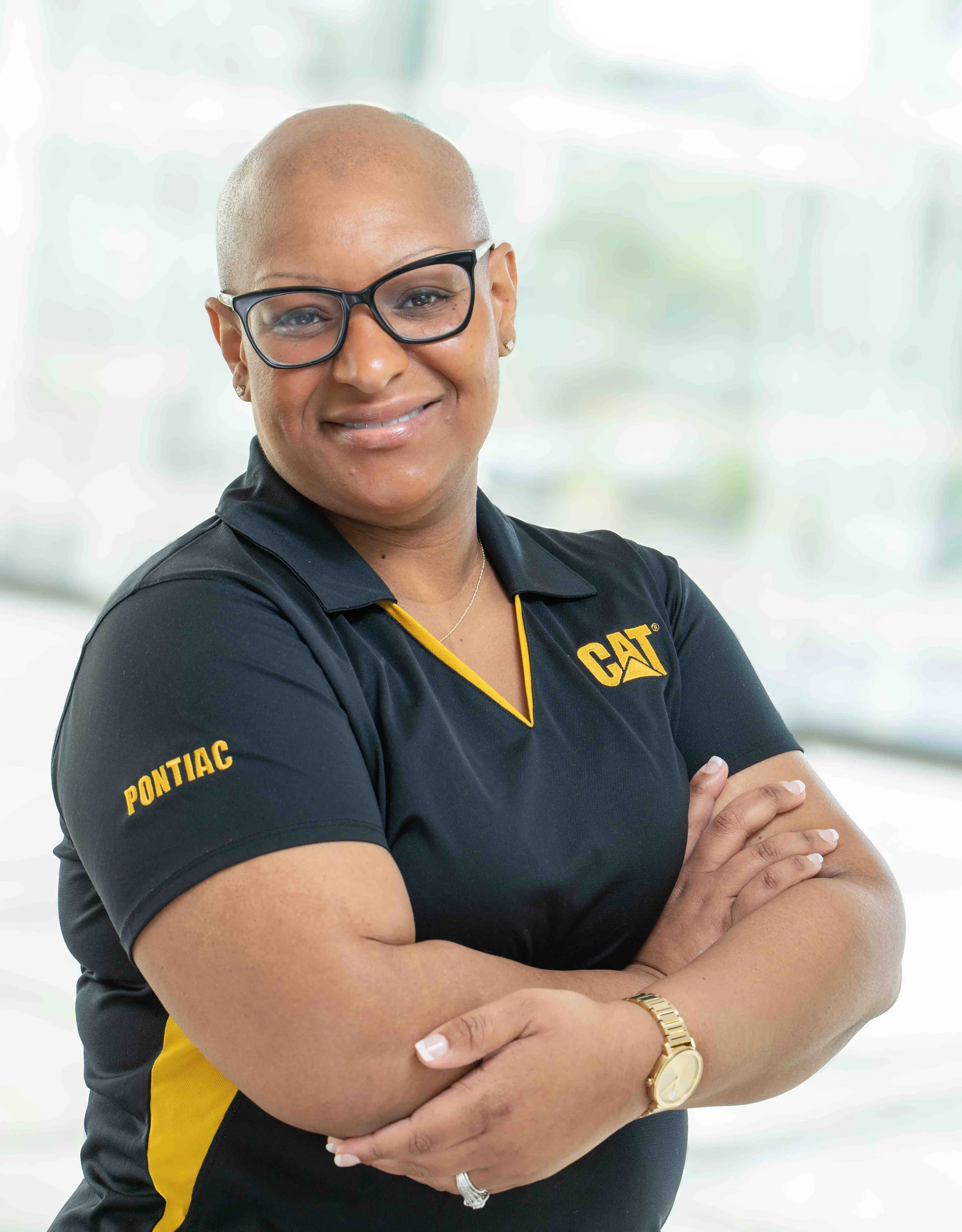 A confident woman with a shaved head, wearing glasses, a black and yellow Polo shirt with 'Pontiac' and 'CAT' logos, crossing her arms and smiling, standing in a bright, blurred background.