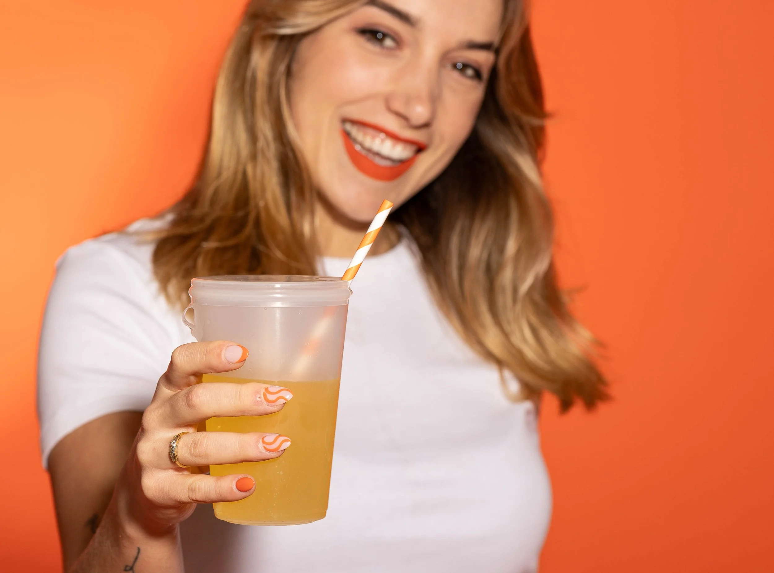 A young woman with brown hair and red lipstick smiling while holding a plastic cup with a yellow beverage and a striped straw, against an orange background.