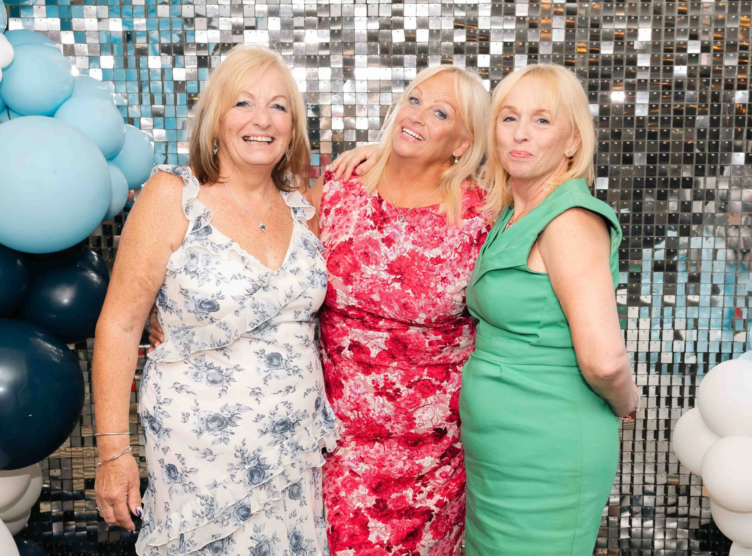 Three women smiling and posing together at a celebration, standing in front of a shiny, reflective backdrop with balloons on either side.