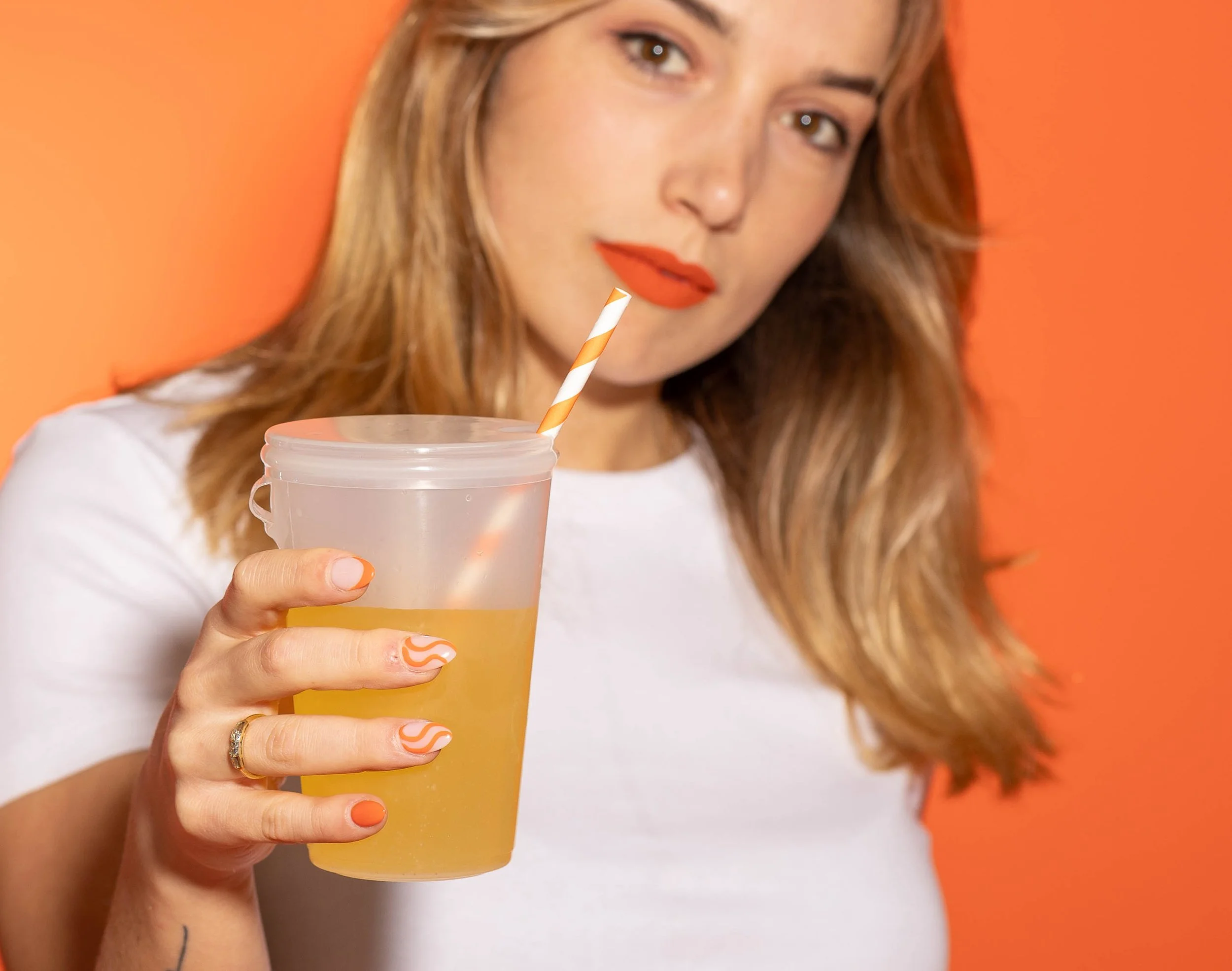 A woman with wavy red hair and orange lipstick holding a transparent cup with a yellow beverage and a striped straw, against an orange background.