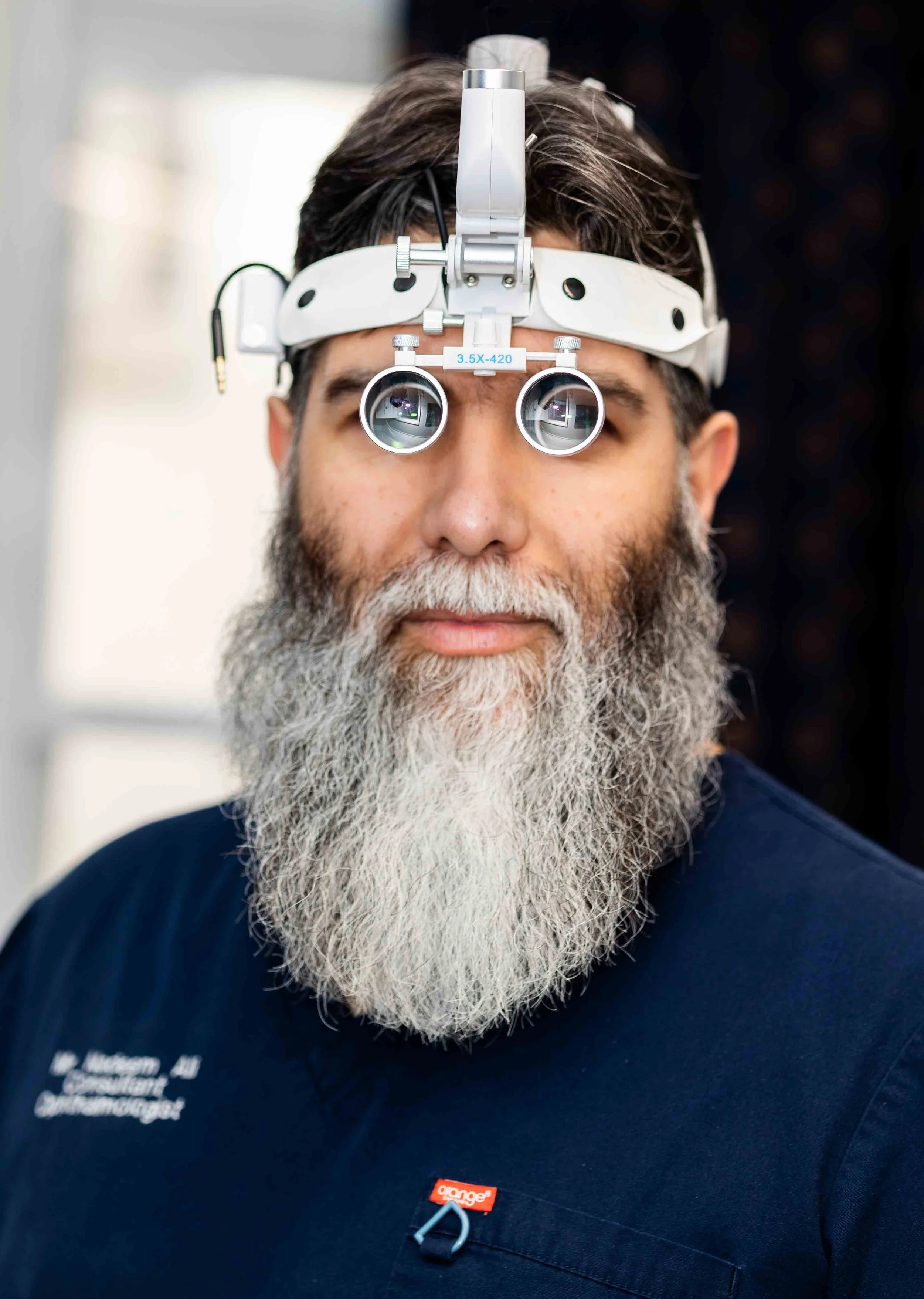 A man with a long gray beard wearing a blue shirt and medical headlamp with magnifying lenses.