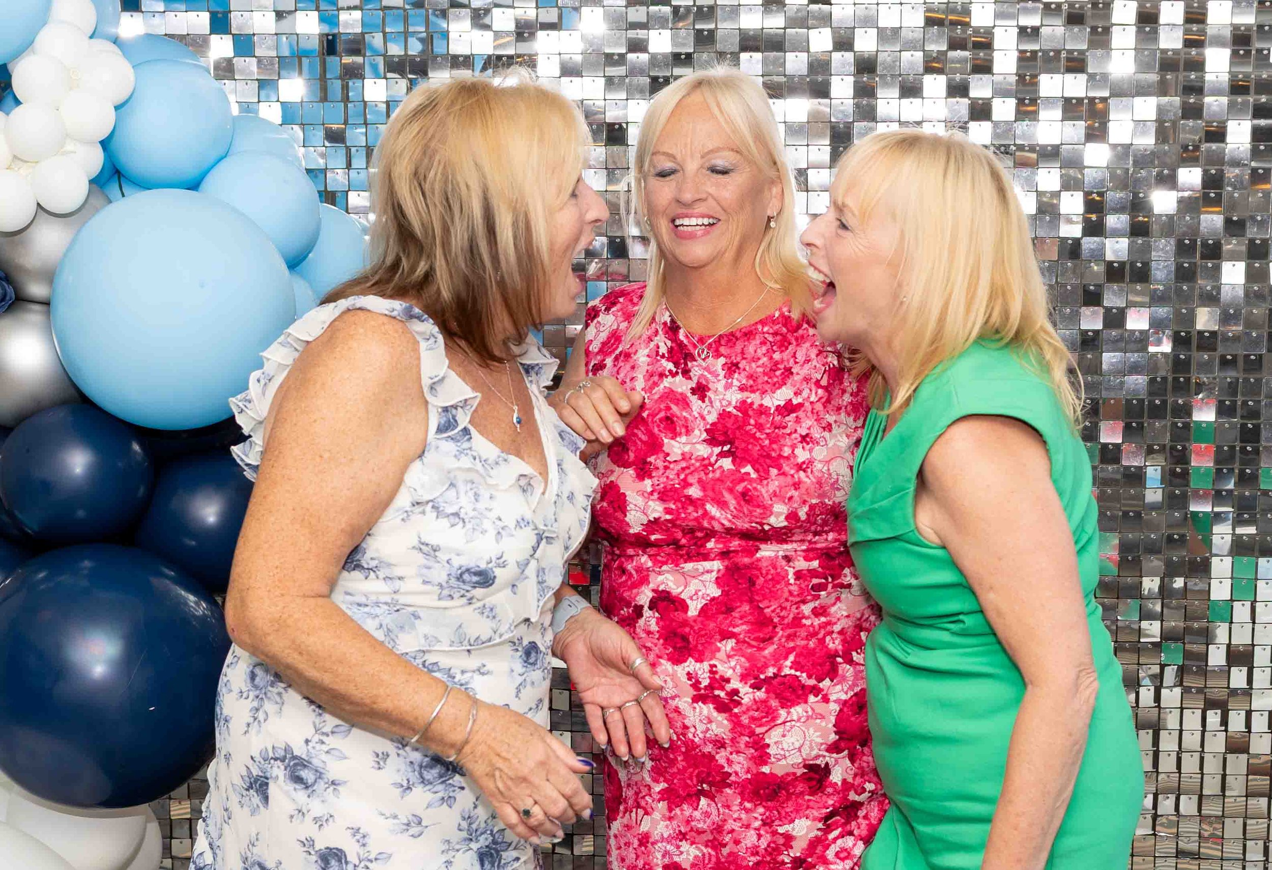 Three women with blonde hair laughing and talking at a celebration, standing in front of a shiny, mosaic tile background and blue and white balloons.