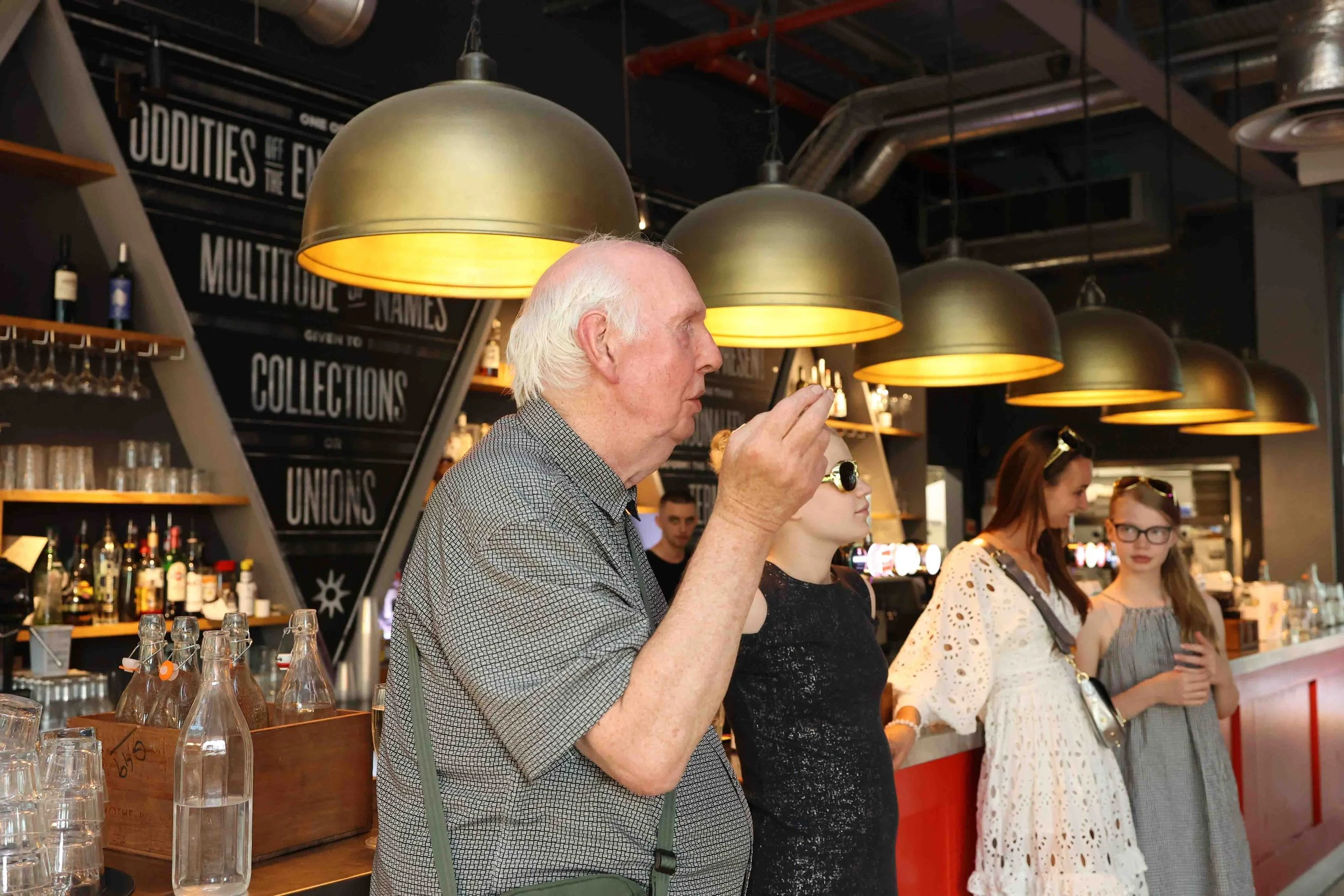 An elderly man with white hair and a gray shirt holding a glass, standing at a bar with three women, in a modern bar or restaurant setting with large gold pendant lamps overhead and black chalkboard walls with text.
