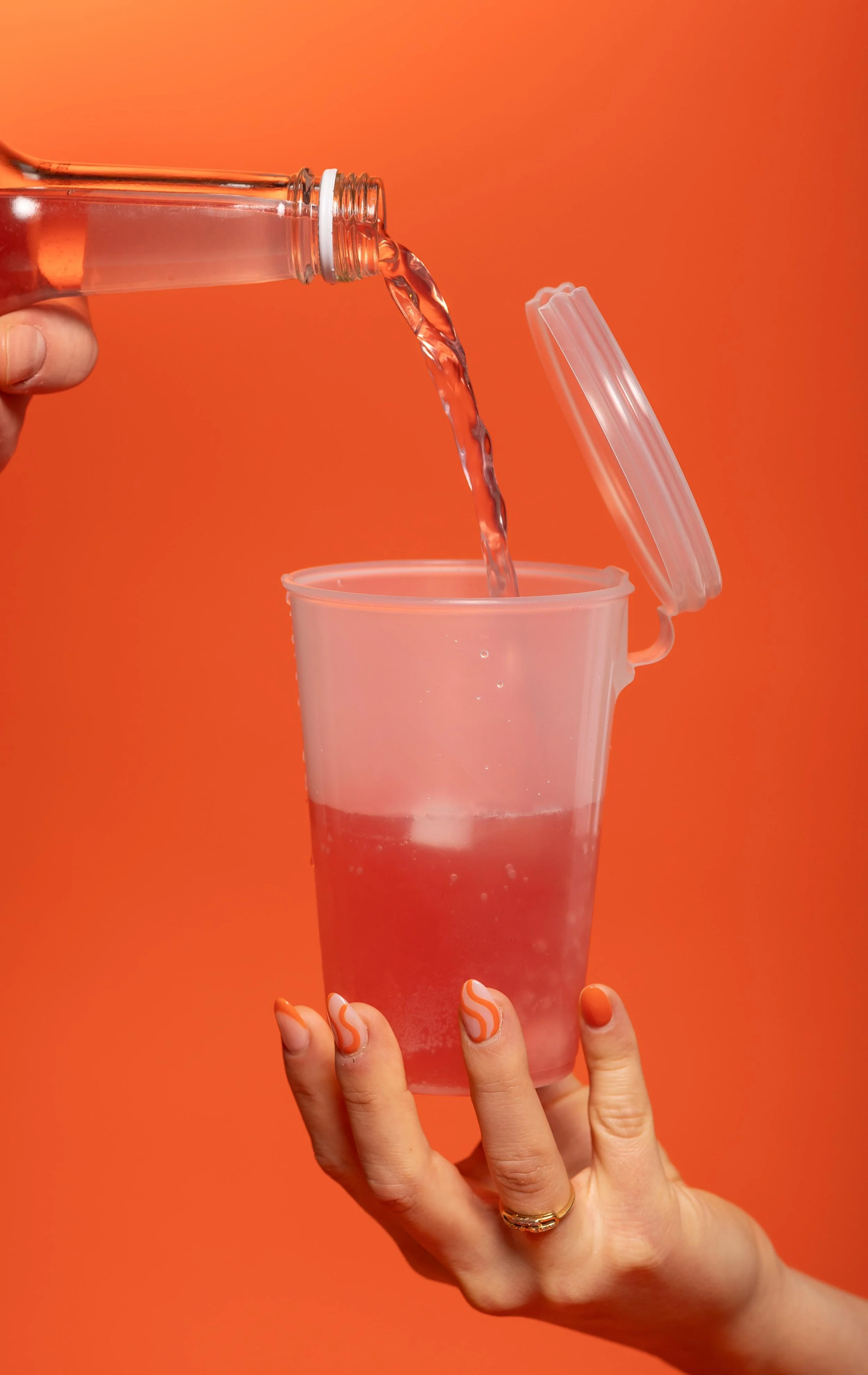 Person holding a pink plastic cup as water is being poured from a glass bottle with orange liquid inside against a vibrant orange background.