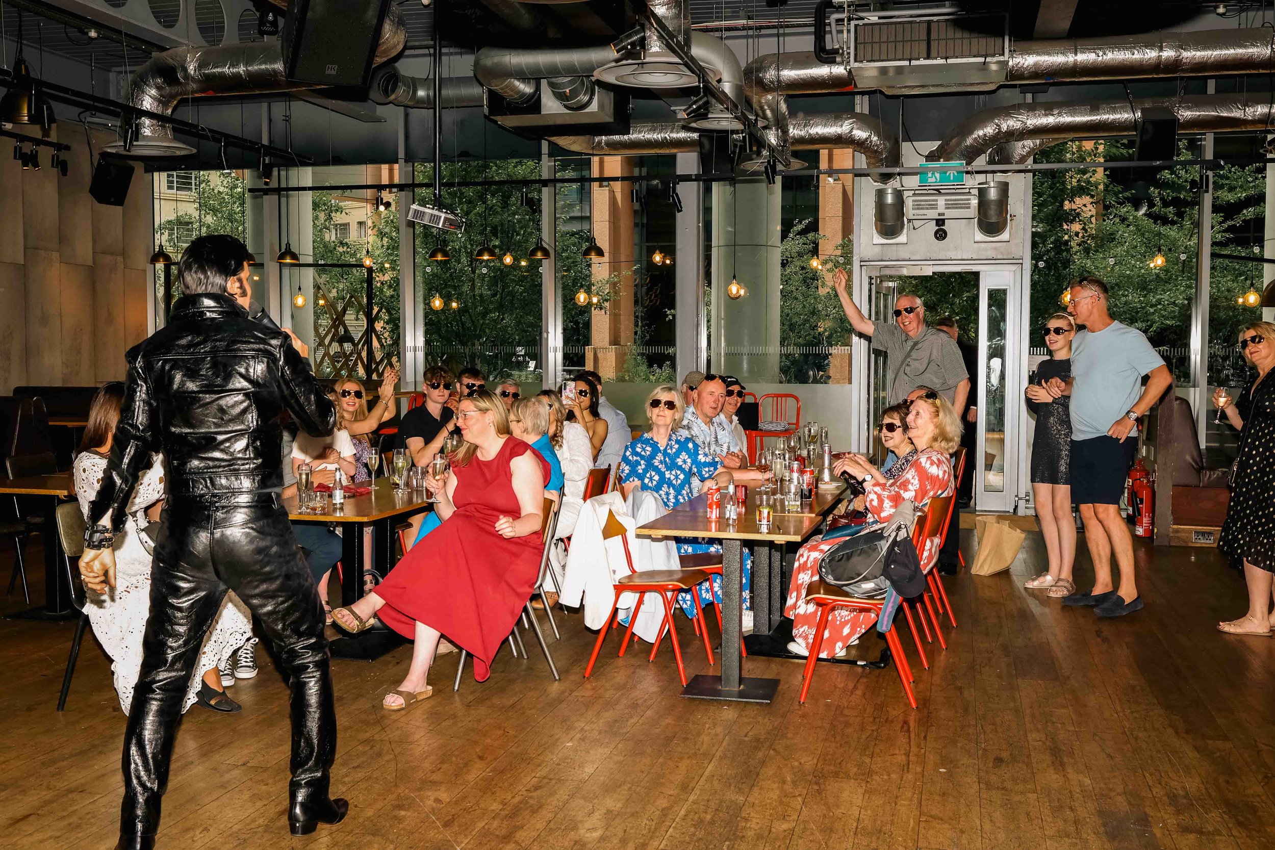 A woman singer performing on stage with an audience seated at tables in a restaurant or event space, with some standing near the back.