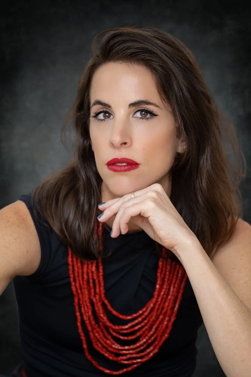 Portrait of a woman with brown hair, wearing red lipstick, a black sleeveless top, and a multi-strand red beaded necklace, posing with her hand near her chin against a gray background.