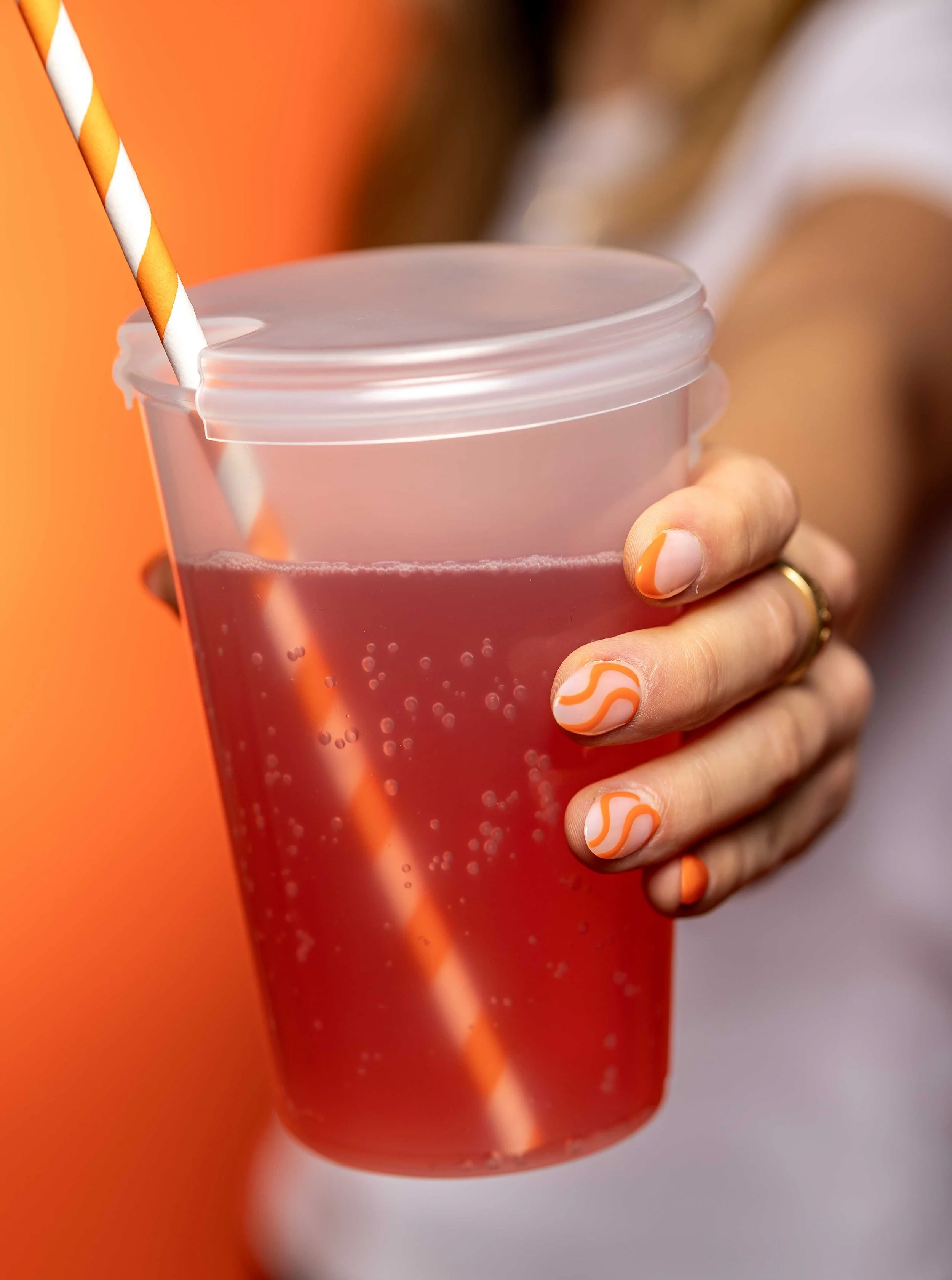 A person holding a transparent cup filled with pink soda, with a striped orange and white paper straw. The person's nails are painted with orange and white wavy designs, and they are wearing a gold ring on their finger.