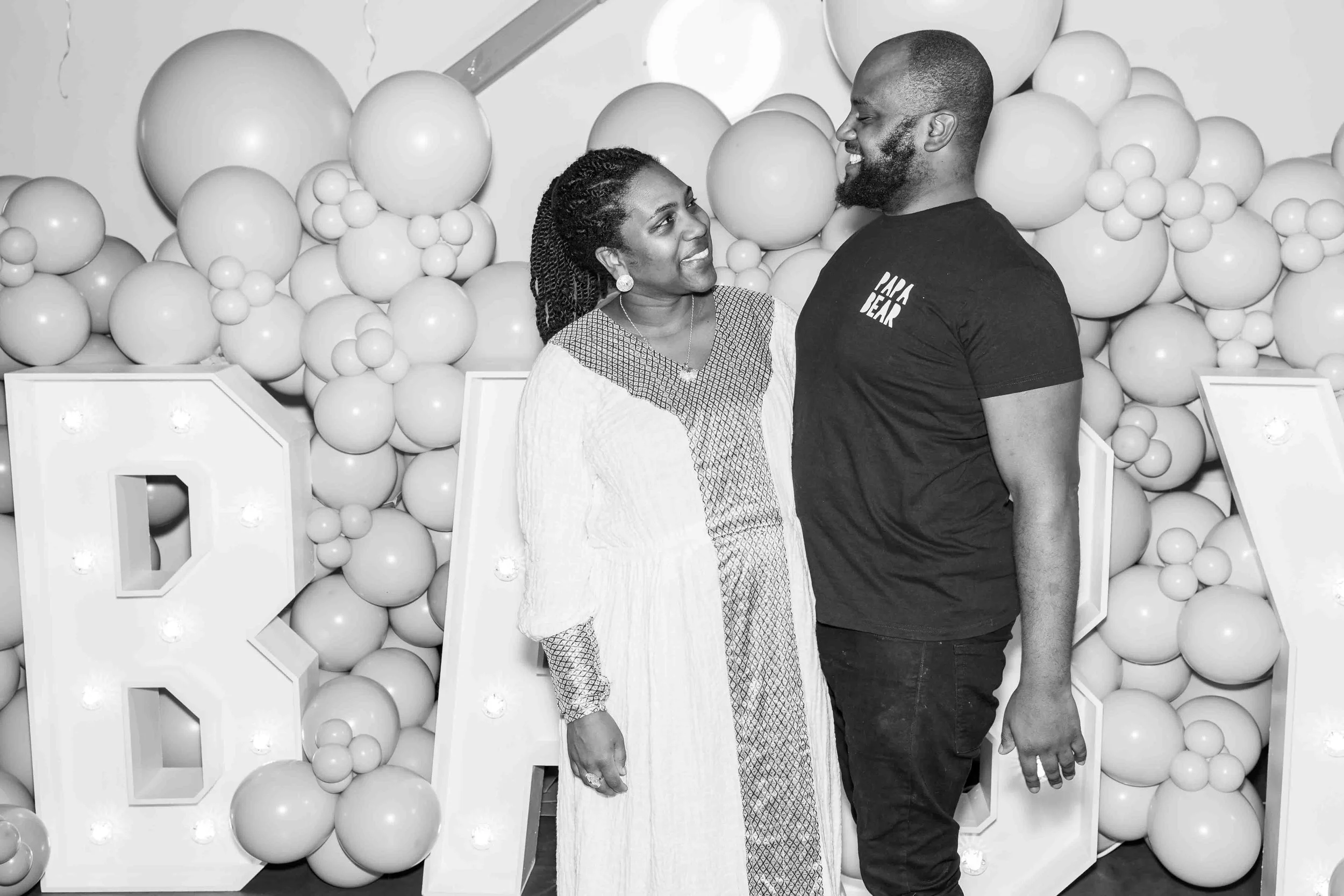 A smiling woman and a man stand close together, looking at each other, in front of a balloon backdrop and large illuminated letters.