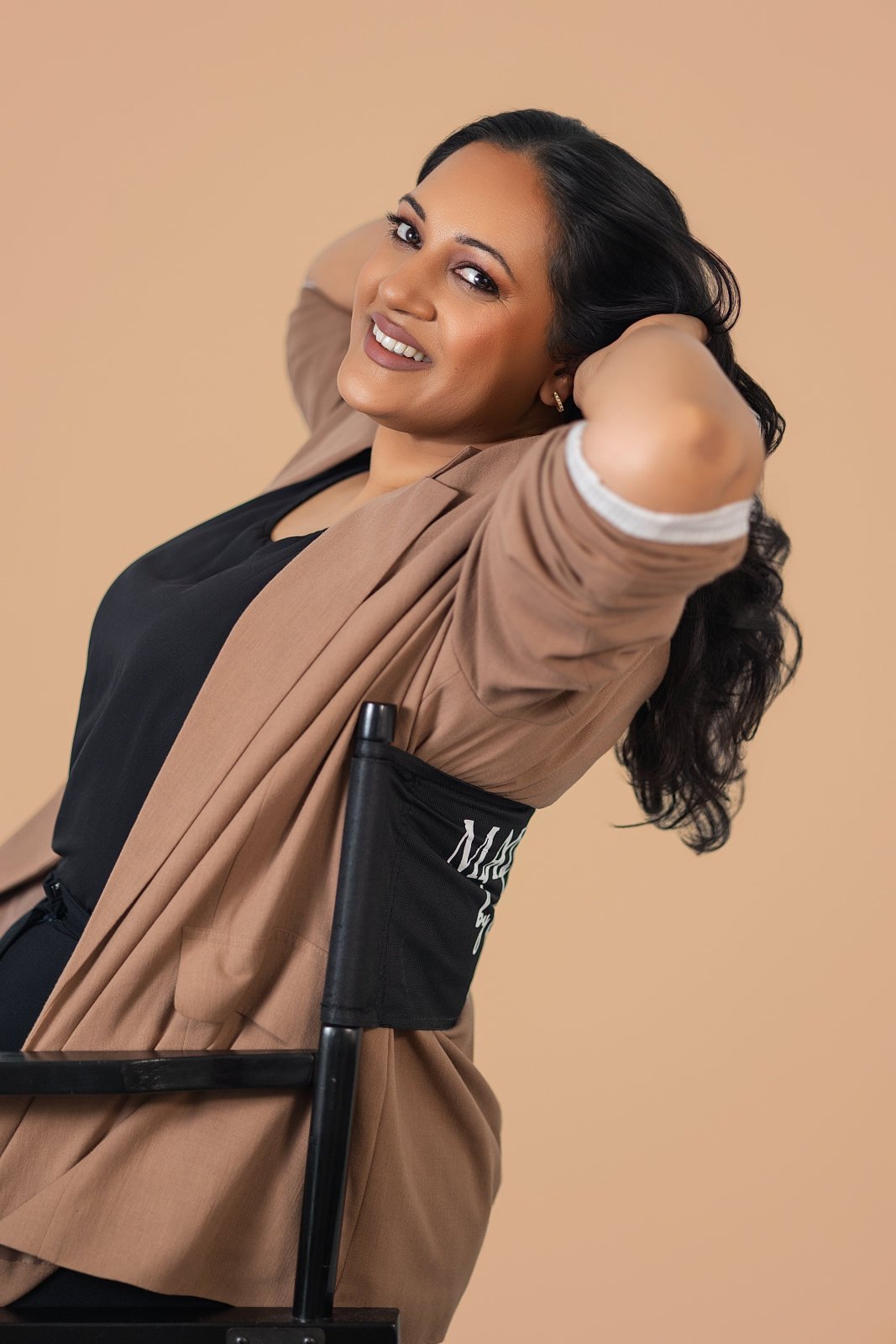 A woman with long black hair is smiling and leaning back in a black director's chair with the background in a beige color.