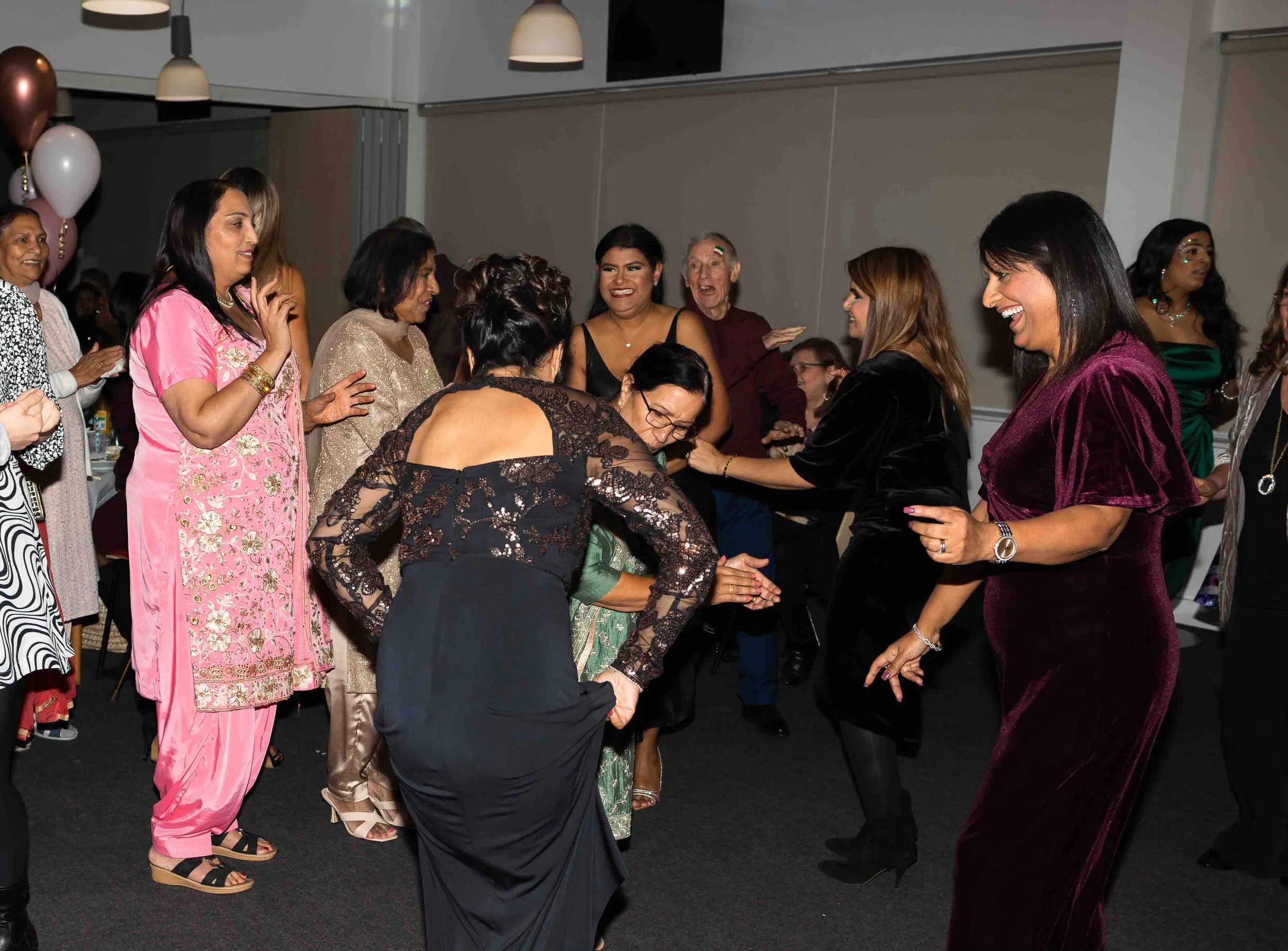 Group of women dancing and laughing at a celebration or party, dressed in colorful and elegant outfits.