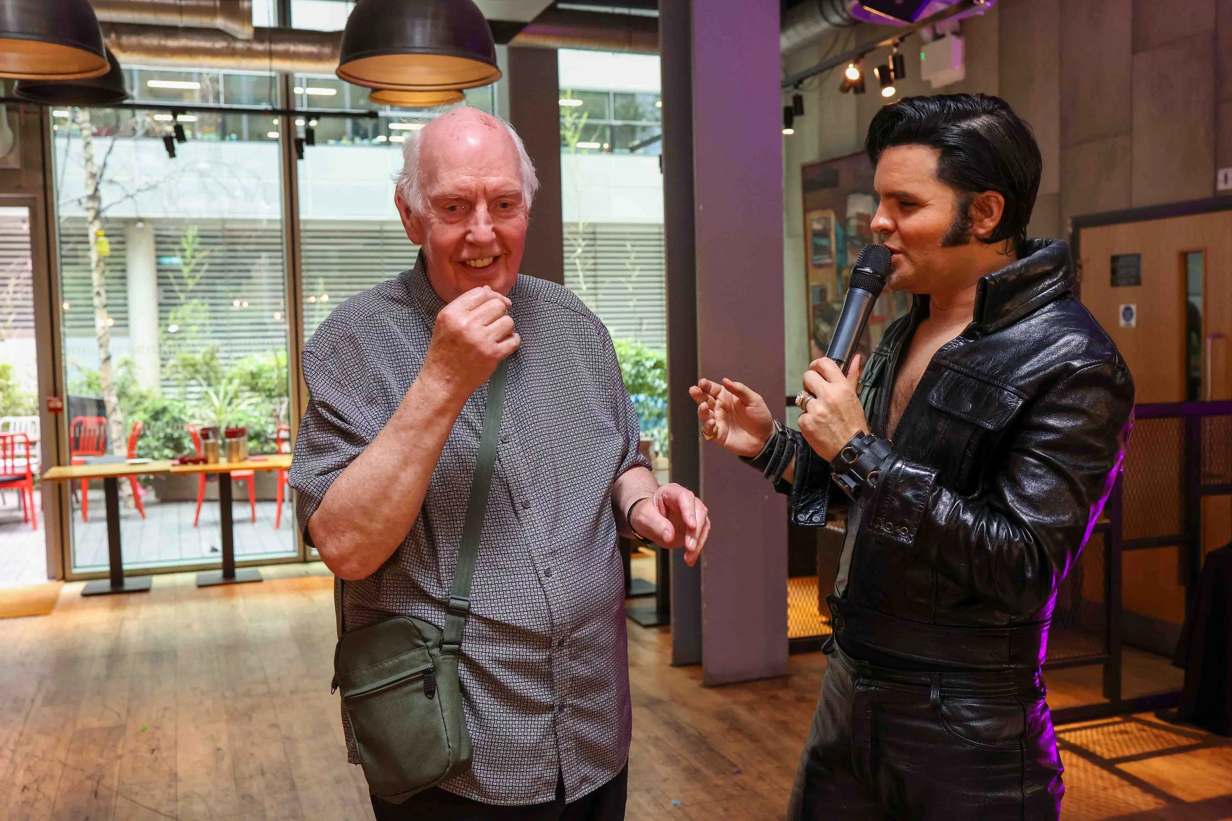 A young man with dark hair, wearing a black leather jacket, speaks into a microphone while an elderly man with white hair, wearing a gray patterned shirt and a small black crossbody bag, listens and smiles in a well-lit indoor space with large window