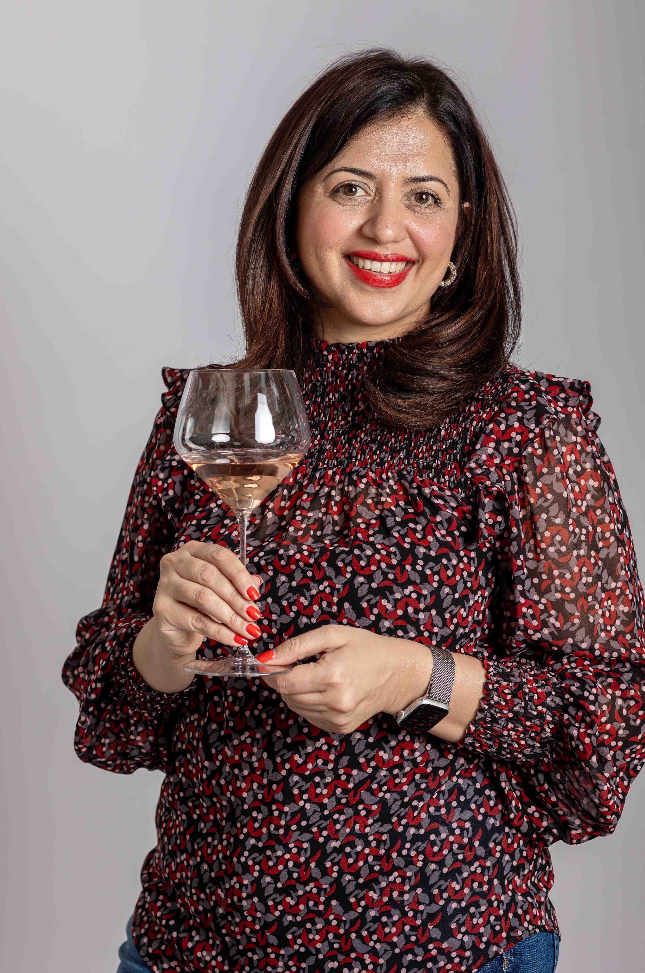 A woman with shoulder-length dark hair and red lipstick, smiling and holding a glass of rosé wine, wearing a patterned blouse with ruffles and a smartwatch on her wrist, against a light gray background.