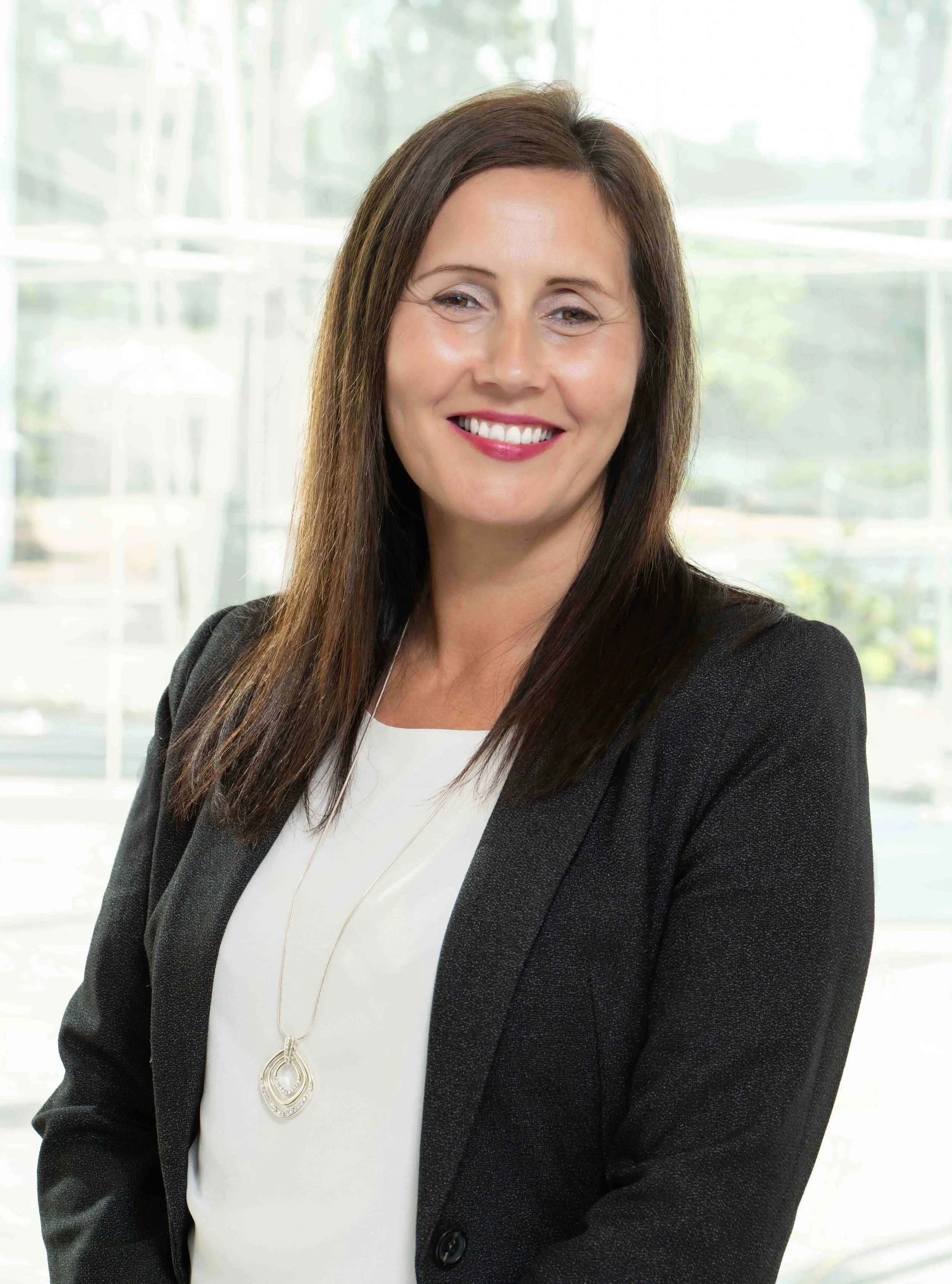 A woman with long brown hair, wearing a black blazer over a white top, smiling in front of a large window.