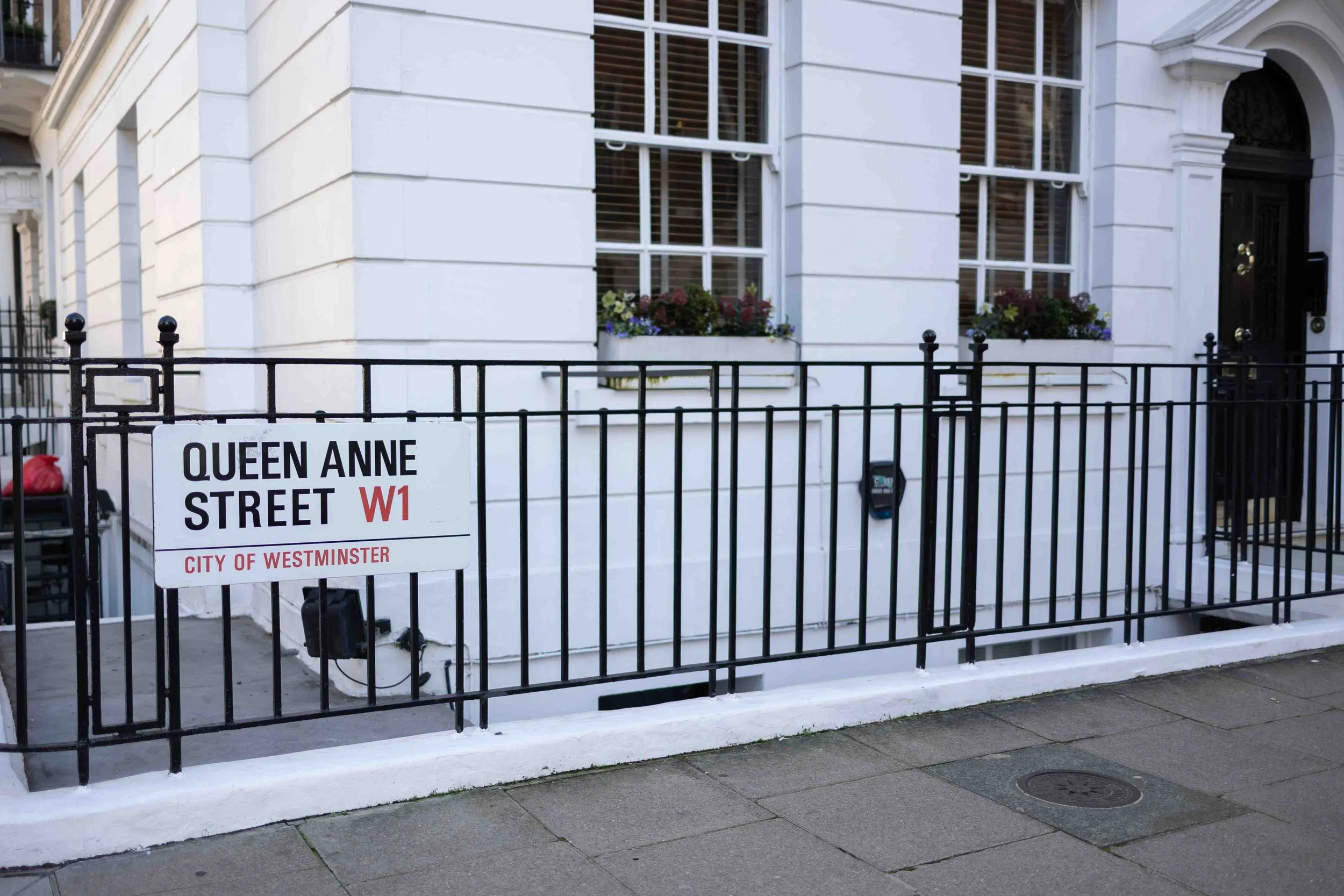 Street sign for Queen Anne Street W1 in Westminster, London, with a black iron fence and a white building with windows and flower boxes in the background.