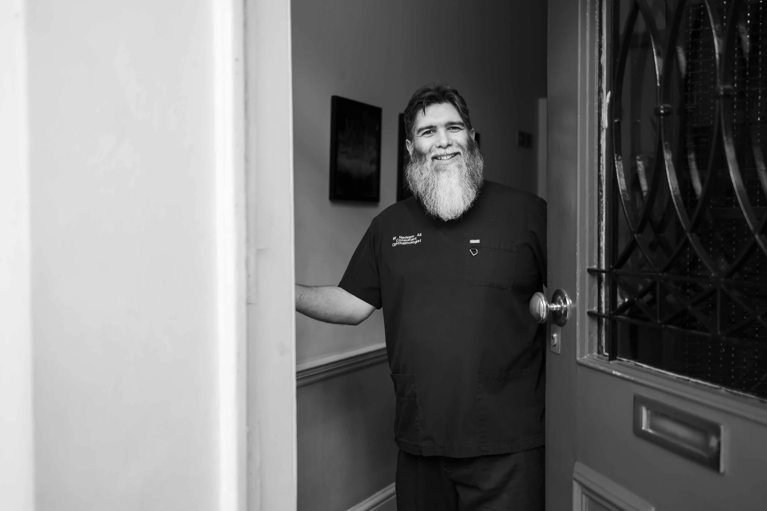 A smiling man with a beard, wearing medical scrubs, standing in a doorway.