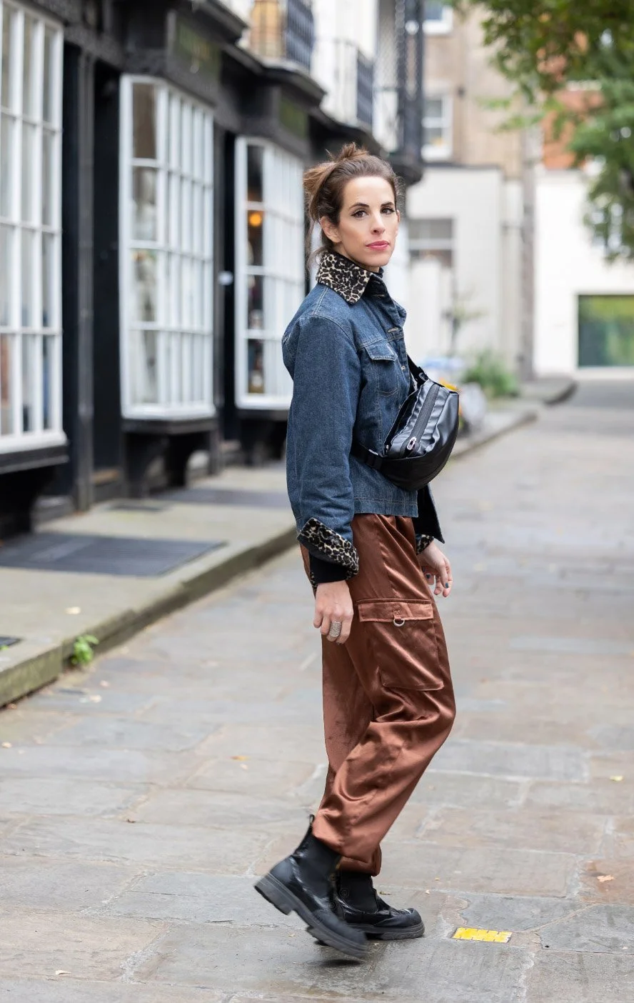 Woman standing on a city sidewalk, wearing a denim jacket with leopard print collar and cuffs, brown satin pants, and black boots, with a black crossbody bag, looking over her shoulder.