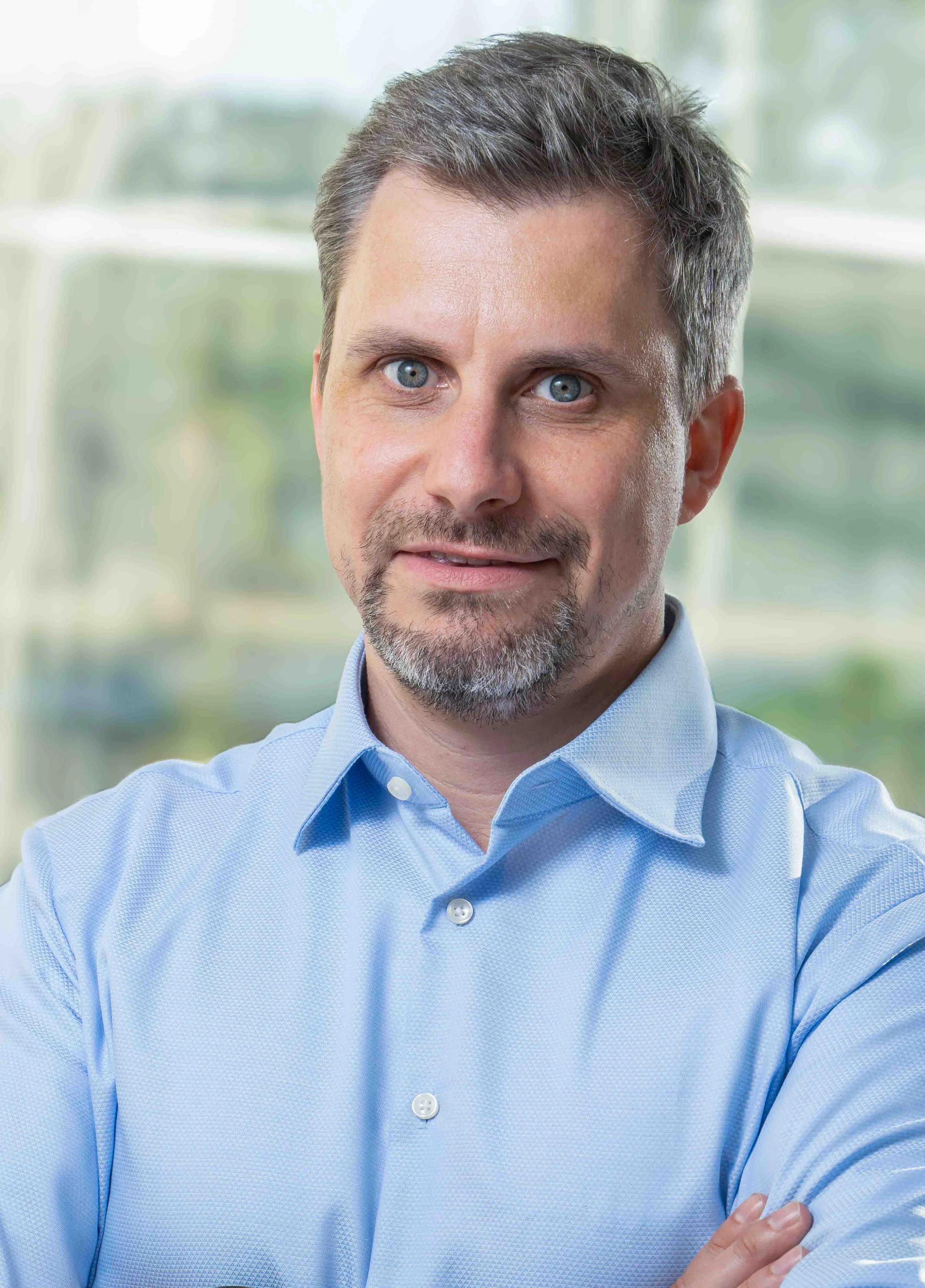 A man with short, styled gray hair and a beard, wearing a light blue dress shirt, standing indoors with a blurred window background.