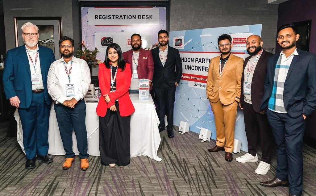 A group of nine professionals standing in front of a registration desk at a conference, with signs indicating a partner conference event, dressed in formal business attire.