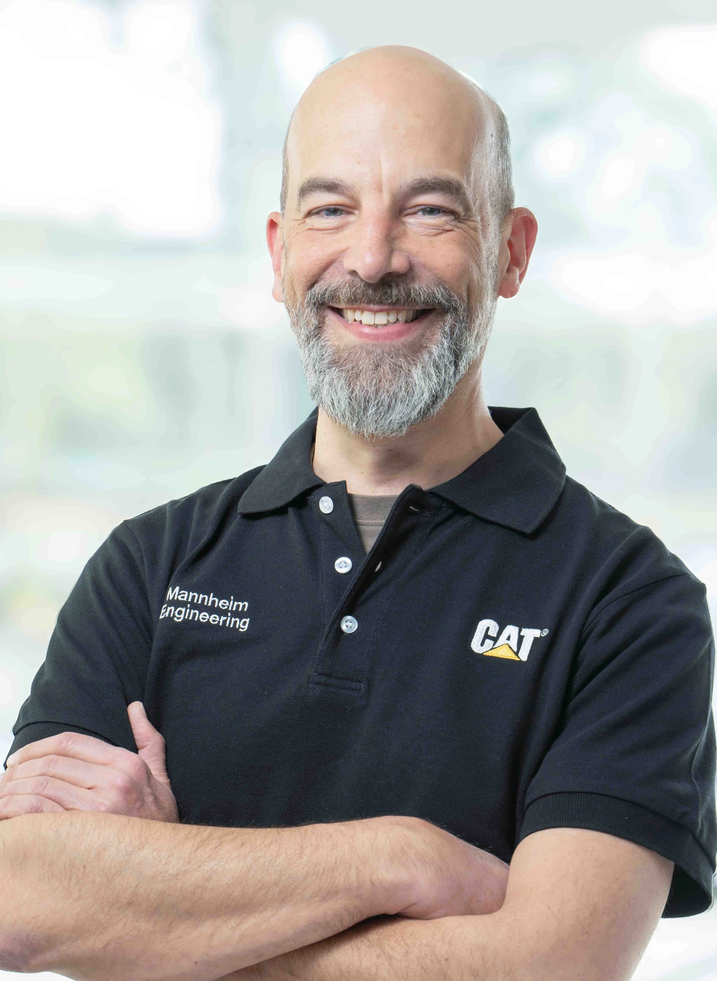 A smiling man with a beard and bald head, wearing a black polo shirt with 'Mannheim Engineering' and 'CAT' logos, standing with arms crossed against a blurred background.