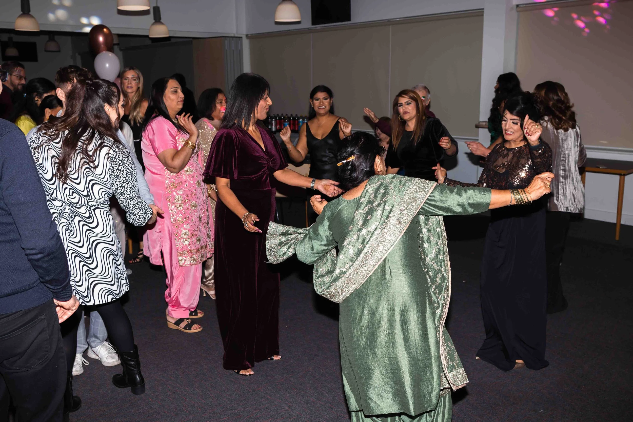 A group of women and one man dancing at an indoor celebration or party, with some wearing traditional South Asian attire and others in modern clothing.