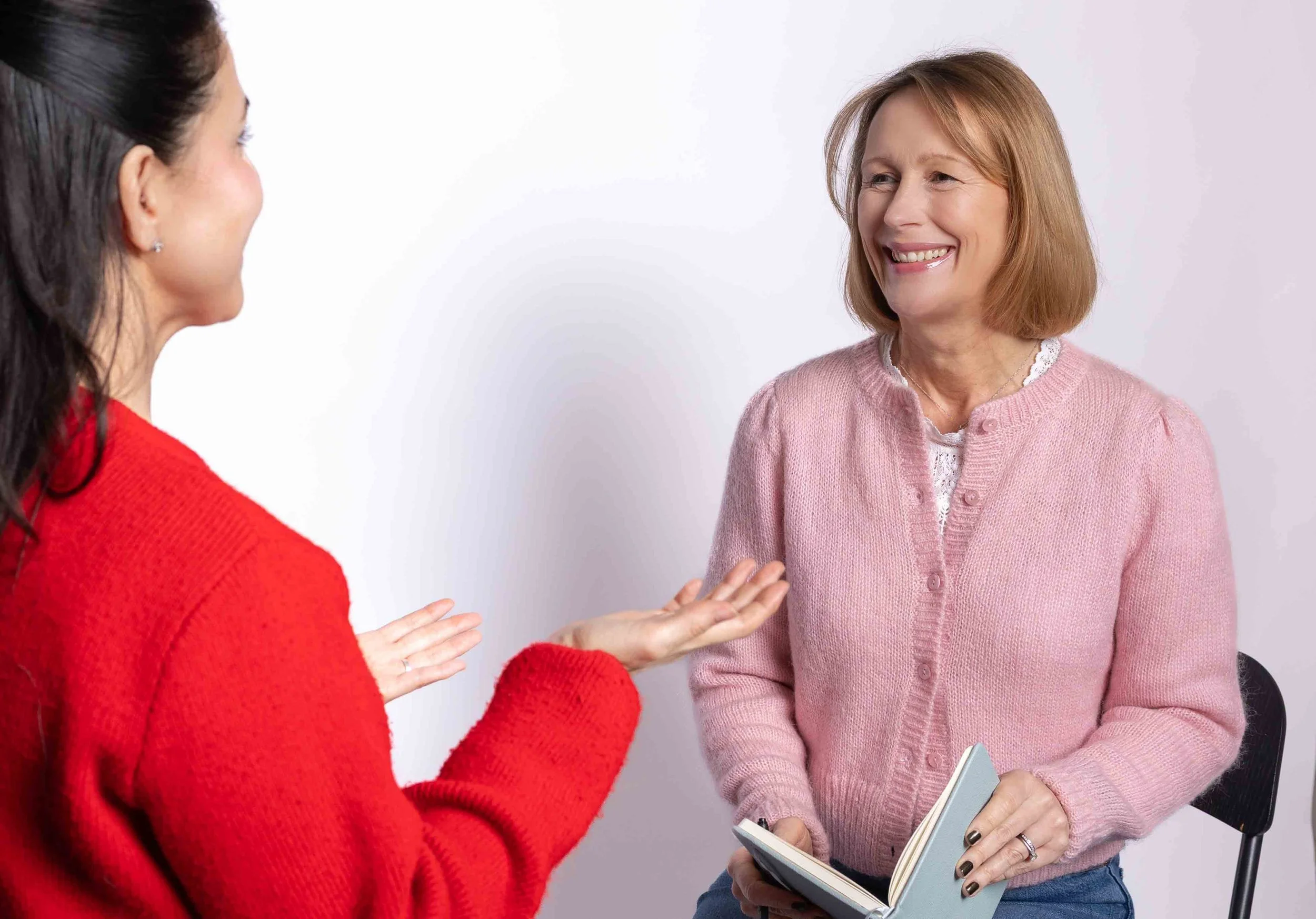 Two women talking, one gesturing with her hands and the other smiling and holding a notebook.