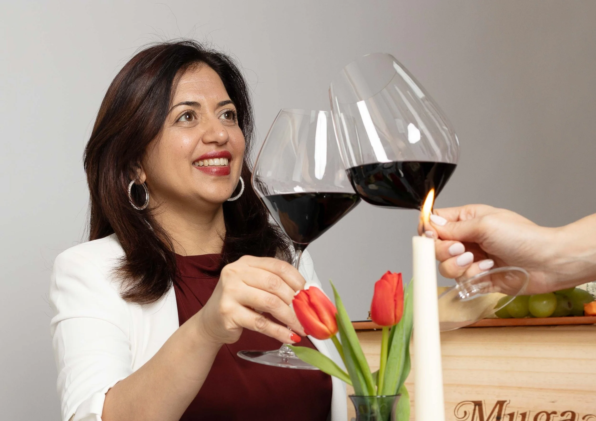 A woman with dark hair and earrings smiles as she holds a glass of red wine during a toast at a celebration. A hand lights a candle on the table, which also has a vase with red tulips and a plate of green grapes.