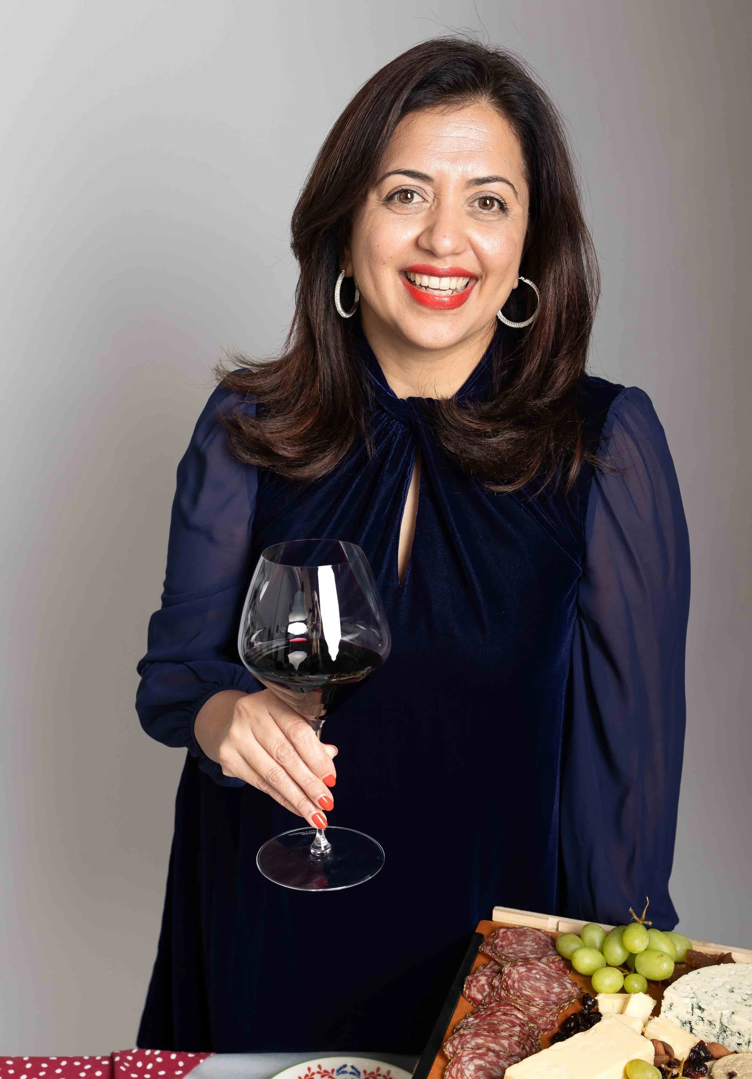 Woman smiling, holding a glass of red wine, with a cheese and meat platter on a table in front of her.