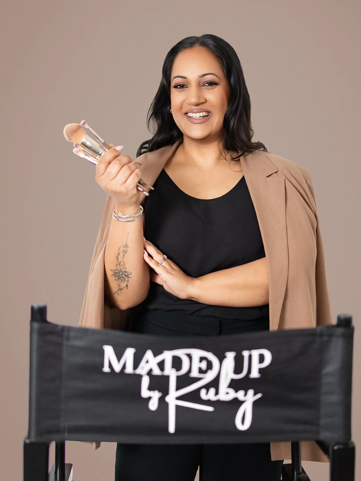 A woman with dark hair, wearing a black shirt and tan blazer, holding makeup brushes, smiling, standing behind a makeup chair with 'MADE UP by Ruby' written on it.