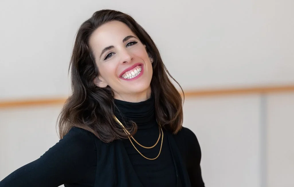 A smiling woman with shoulder-length dark brown hair, wearing a black turtleneck sweater, layered gold necklaces, and makeup, standing against a plain background.