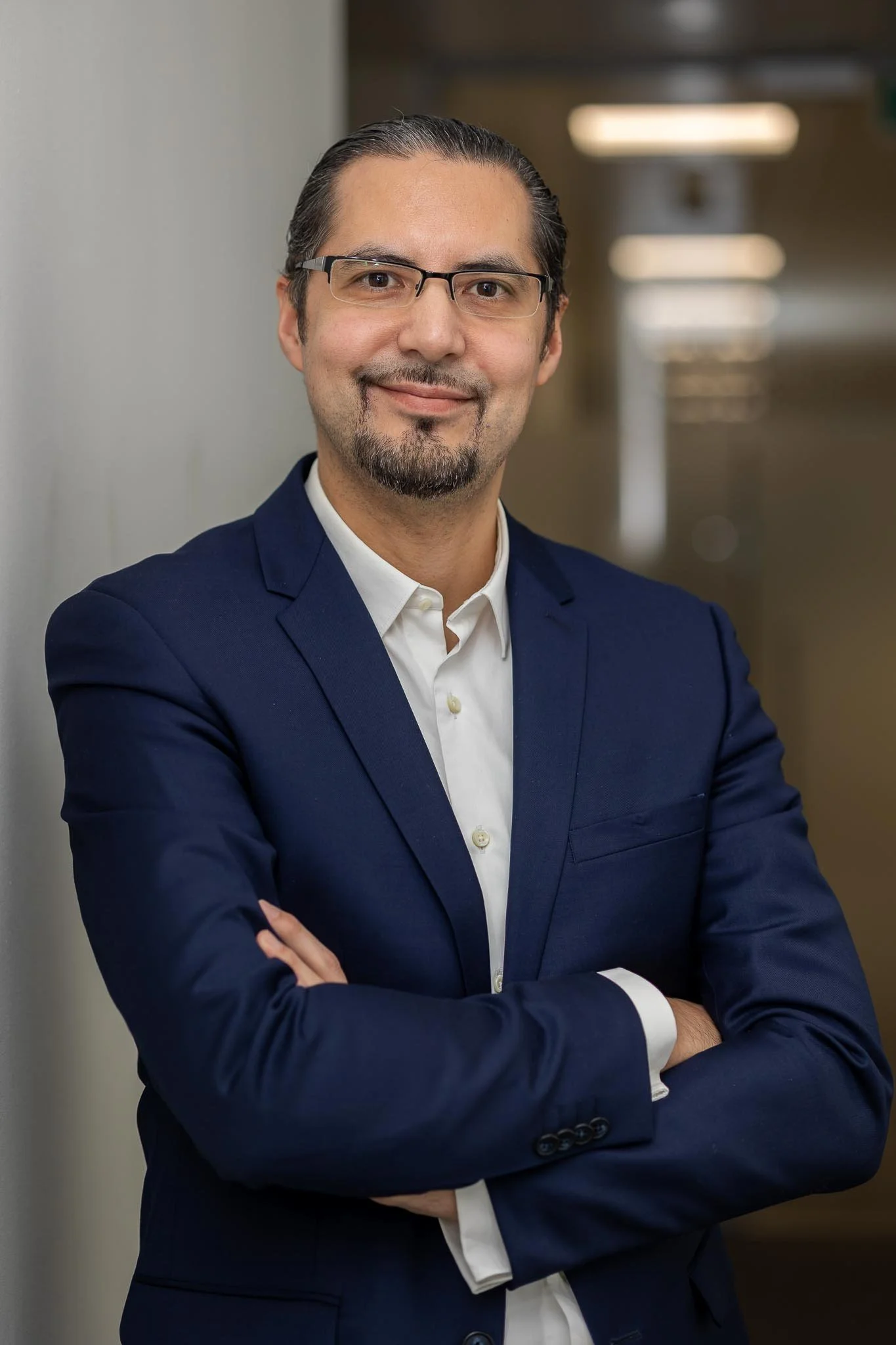 A confident man with glasses, a goatee, and neatly styled hair, wearing a navy blue suit and white shirt, standing with arms crossed in a professional setting.
