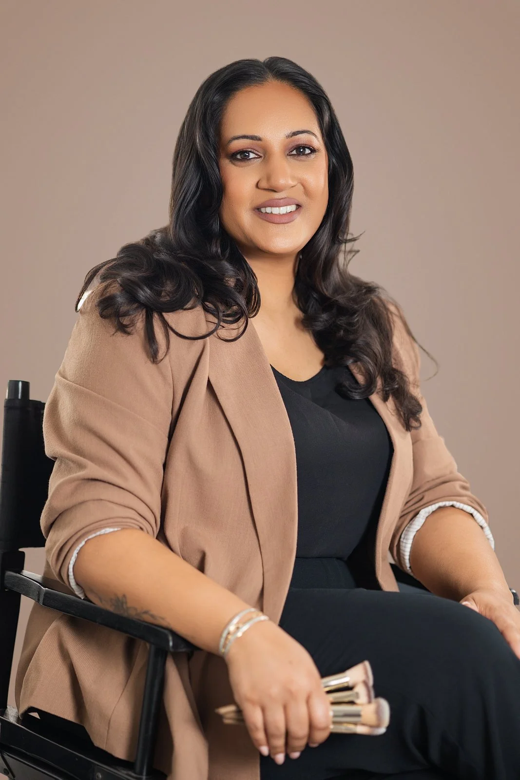 A woman with long dark hair, wearing a black top and a tan blazer, sitting in a wheelchair, smiling at the camera.